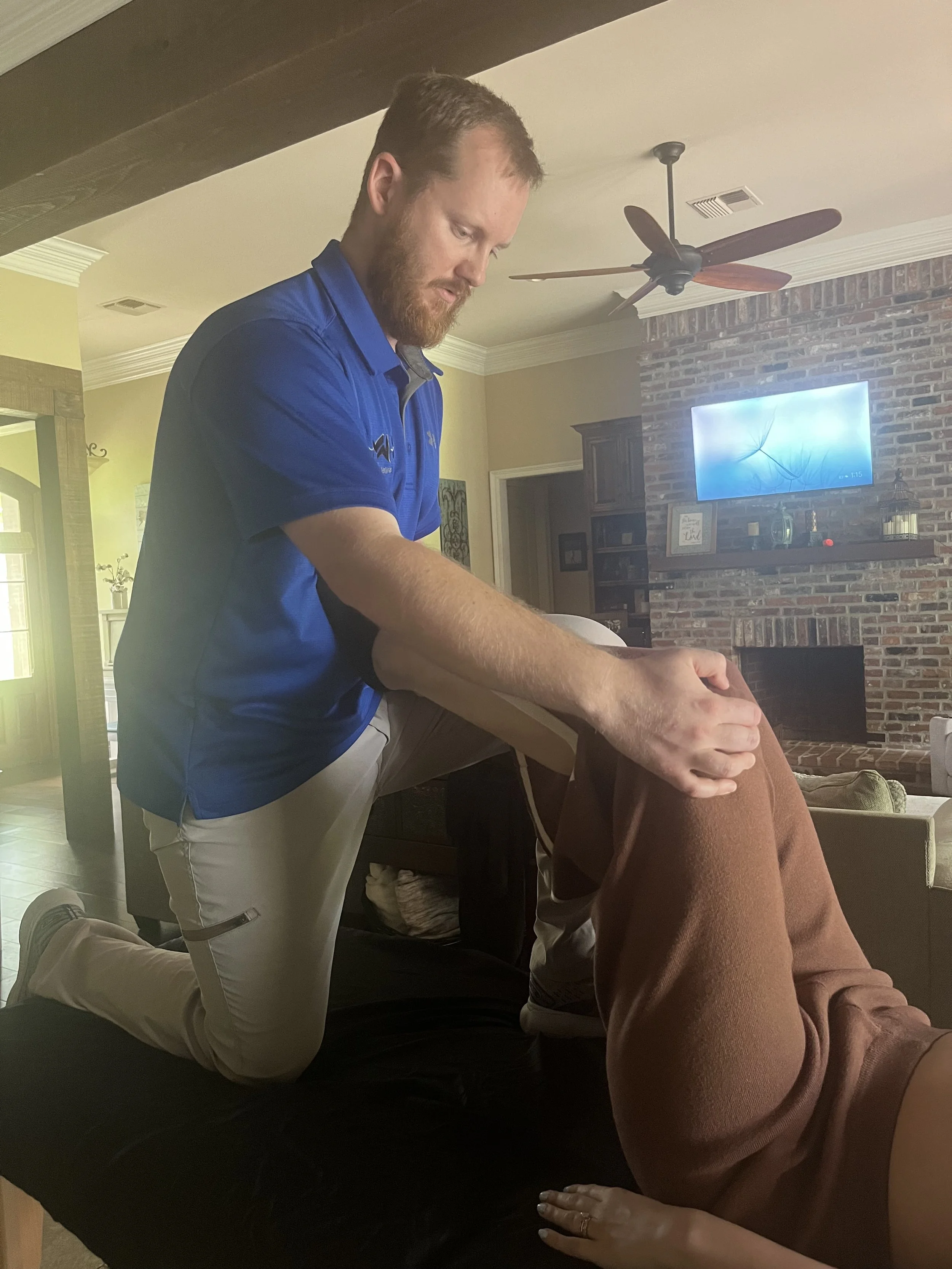 A man is kneeling on a massage table stretching out a person lying down. The man is wearing a blue shirt and beige pants, and the room has a brick fireplace and a ceiling fan.