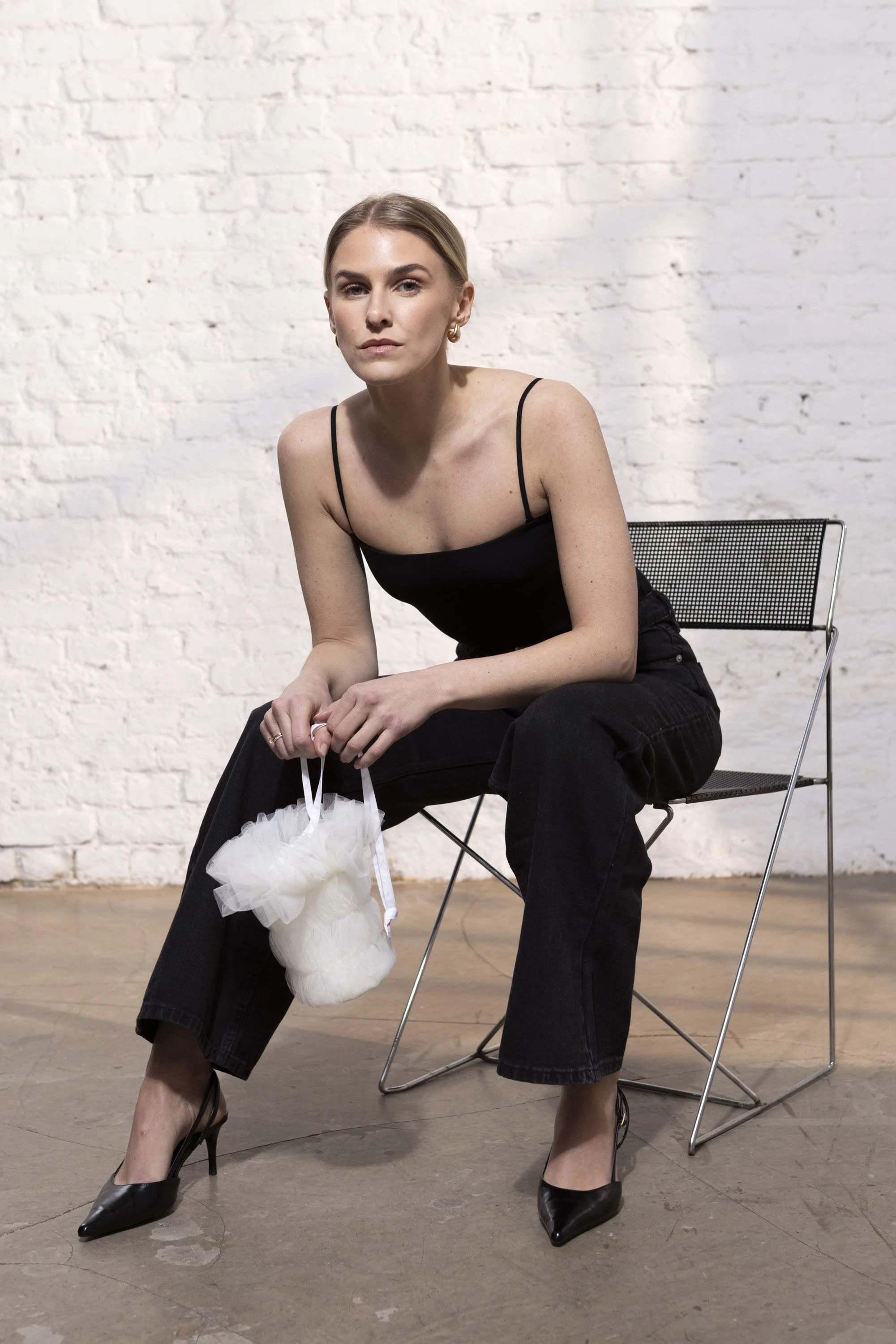A woman holding a unique bridal bag, costume made, with blonde hair in a ponytail, wearing a black spaghetti strap top, black jeans, and black high heels, sits on a metal wire chair holding a small white purse, against a white brick wall.
