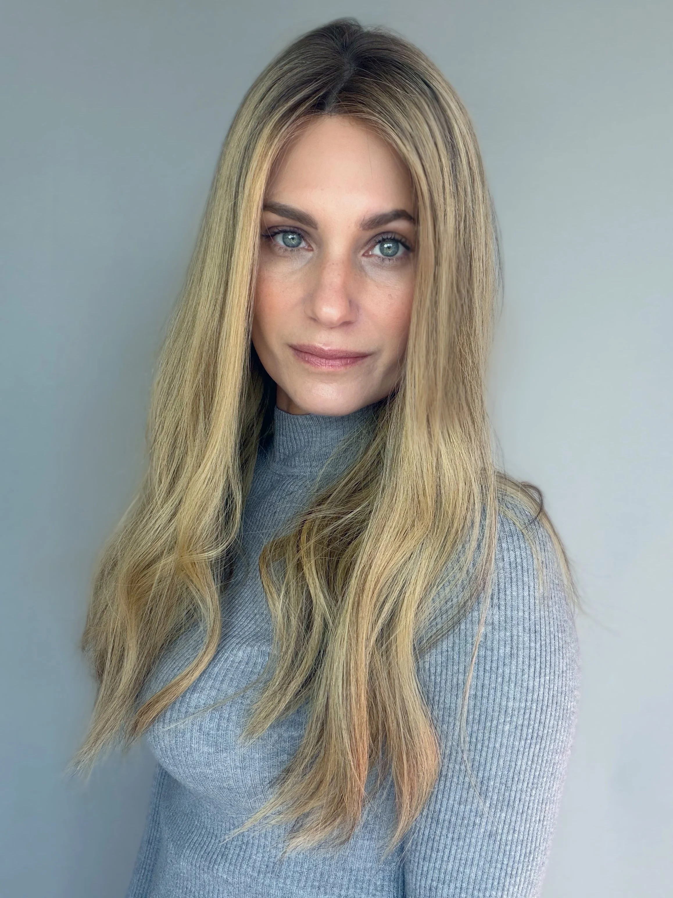 A woman with long blonde hair, blue eyes, and fair skin wearing a gray turtleneck sweater, standing against a plain light gray wall.