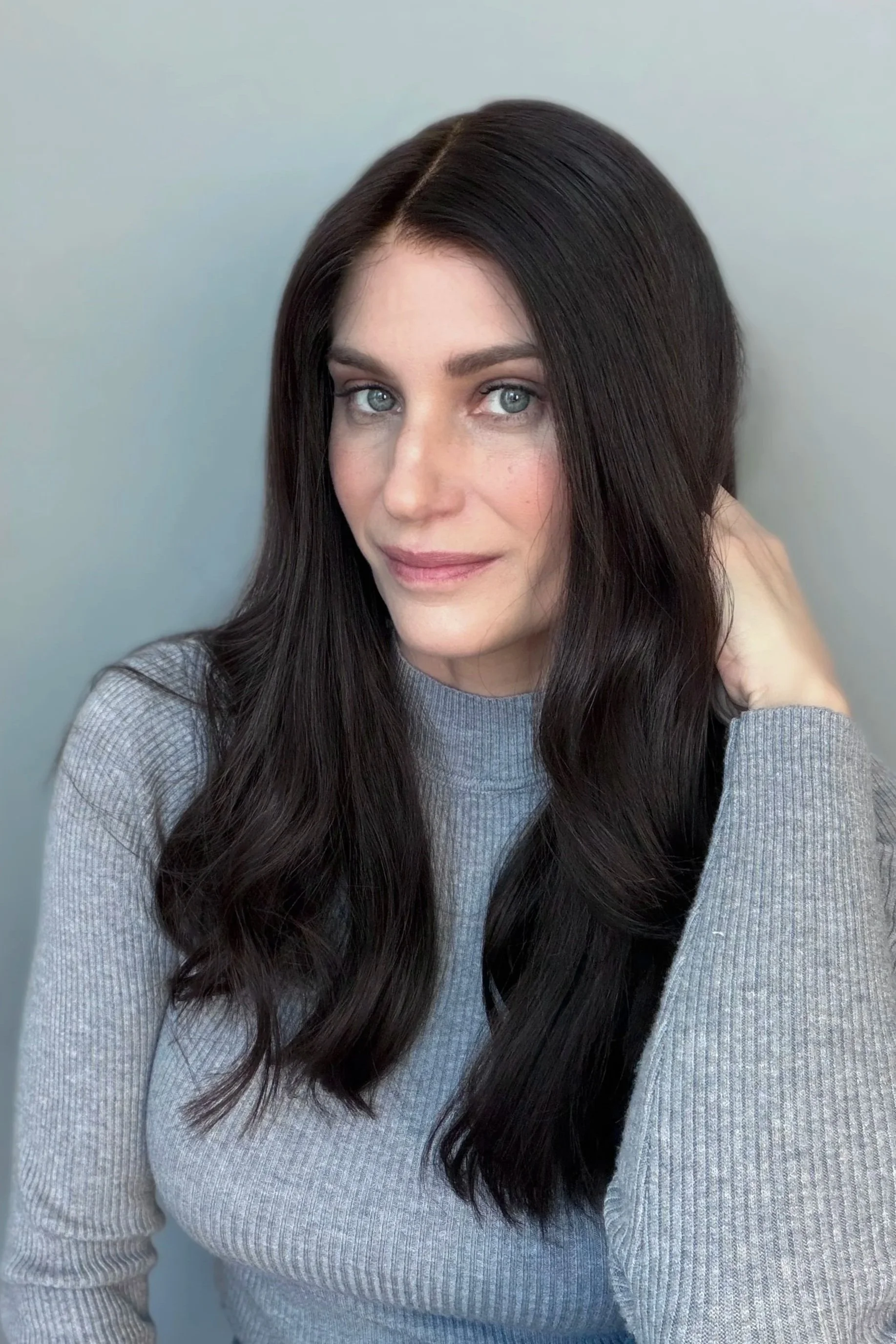A woman with long, dark hair and blue eyes, wearing a light gray knit sweater, posing against a plain light blue background.