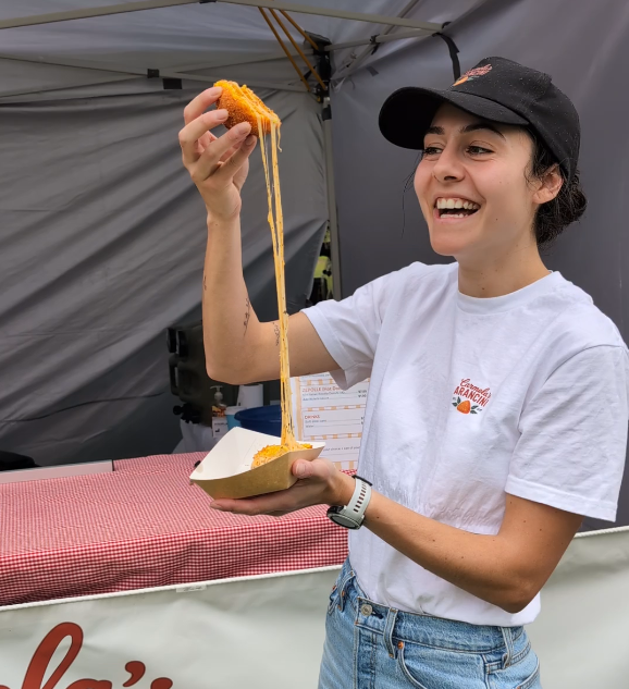 A Co-owner holding an arancini with stretched cheese, smiling at an outdoor food stand.