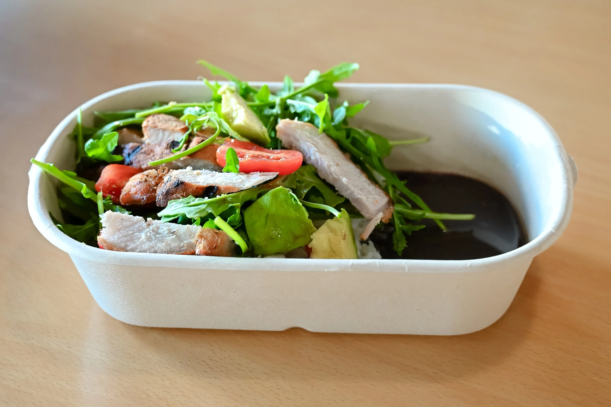 A bowl of salad containing mixed greens, cherry tomatoes, shredded chicken, avocado, and sliced turkey or roast beef with a dark dressing.