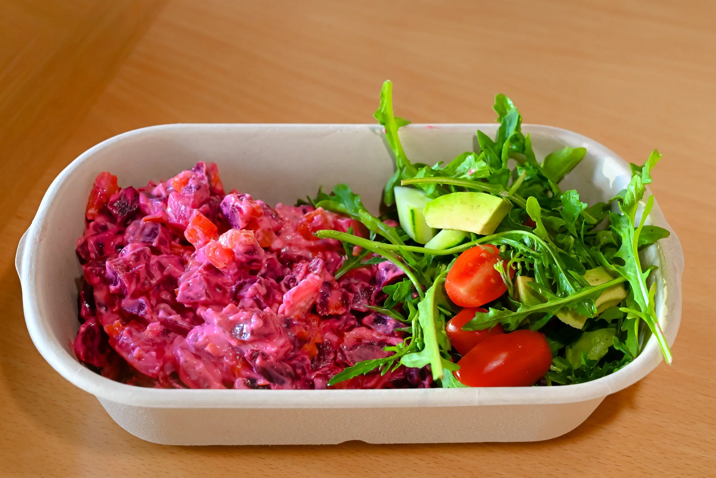 A rectangular container with a salad of green lettuce, cherry tomatoes, slices of cucumber, and a side of beetroot salad with a creamy dressing.