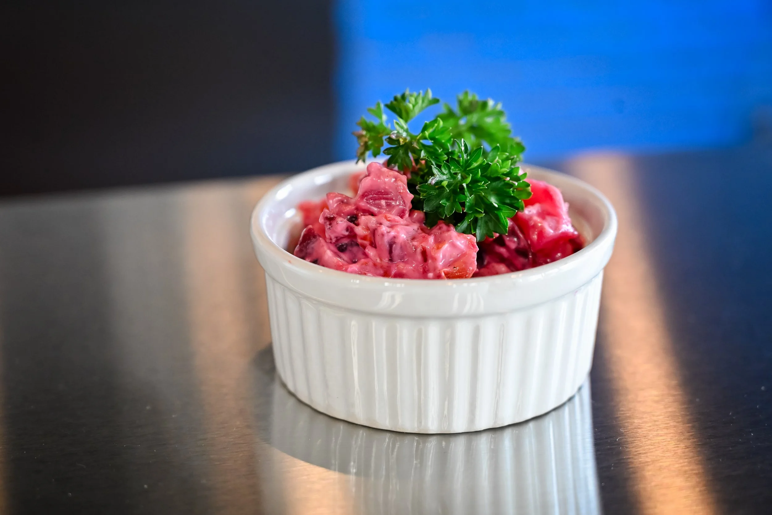 A white ramekin with a textured exterior, filled with a pinkish salad that appears to contain chunks of tuna or chicken and mayonnaise or dressing, topped with a sprig of fresh parsley, on a reflective metal surface.