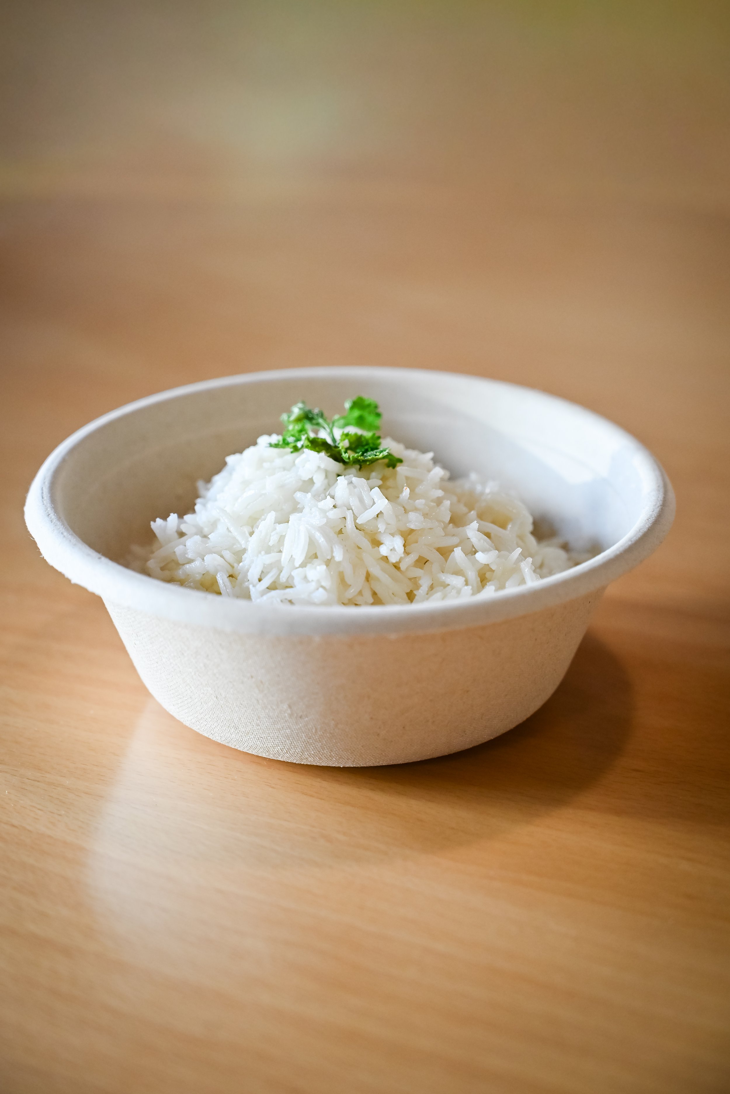 A white bowl filled with cooked white rice, garnished with a small green herb, placed on a light wooden surface.