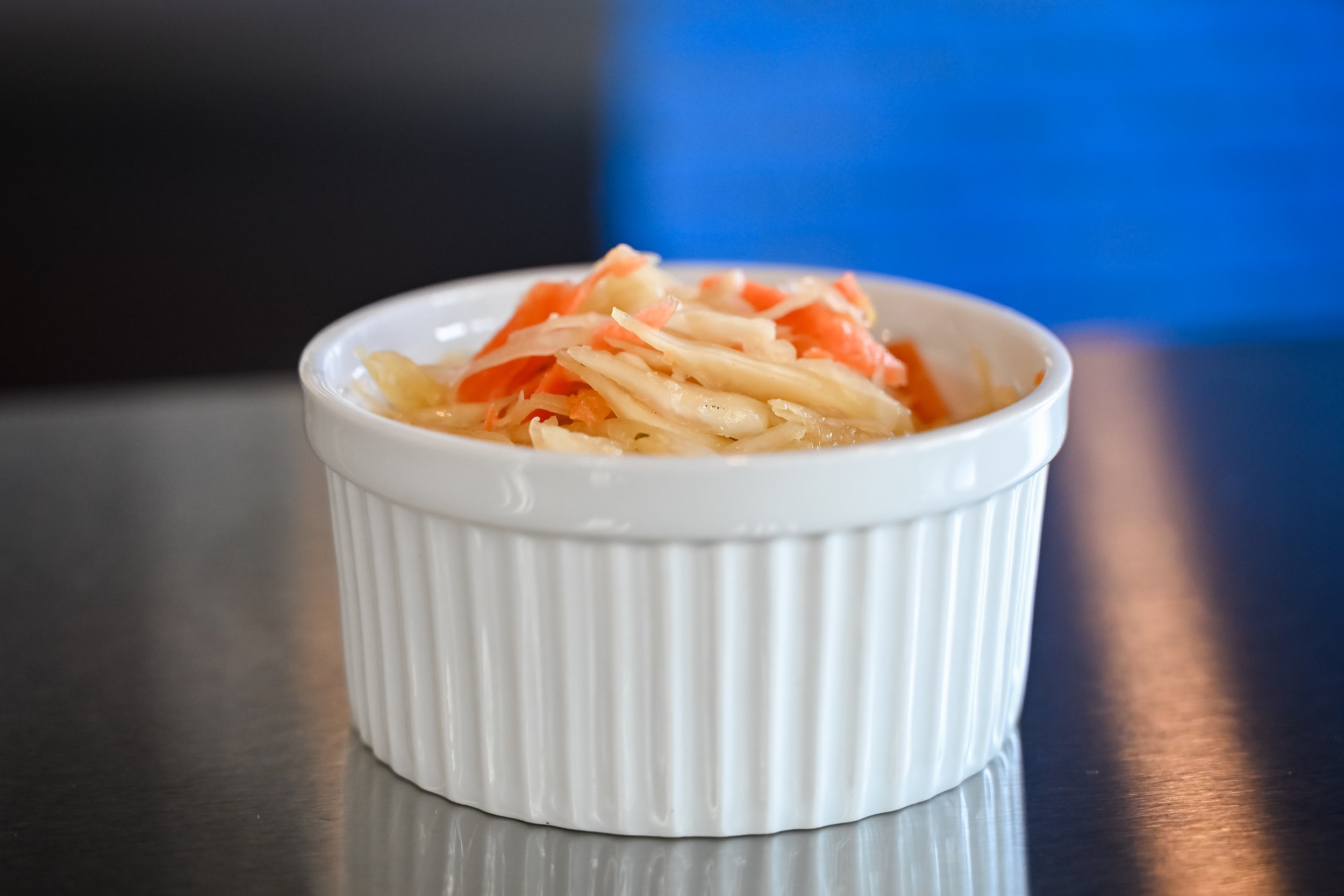 A small white ramekin filled with coleslaw, containing shredded cabbage and carrots, is placed on a dark reflective surface with a blurry blue background.