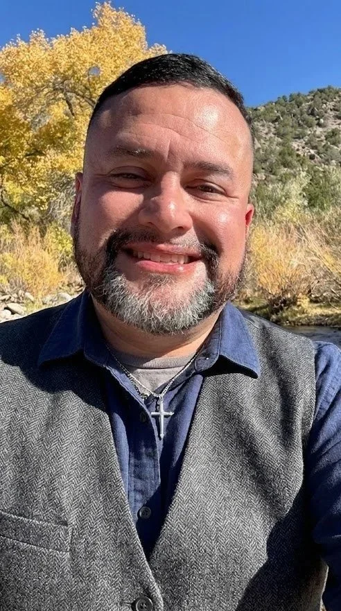 A man smiling outdoors with yellow trees and a hillside in the background, wearing a blue shirt, gray vest, and a silver cross necklace.