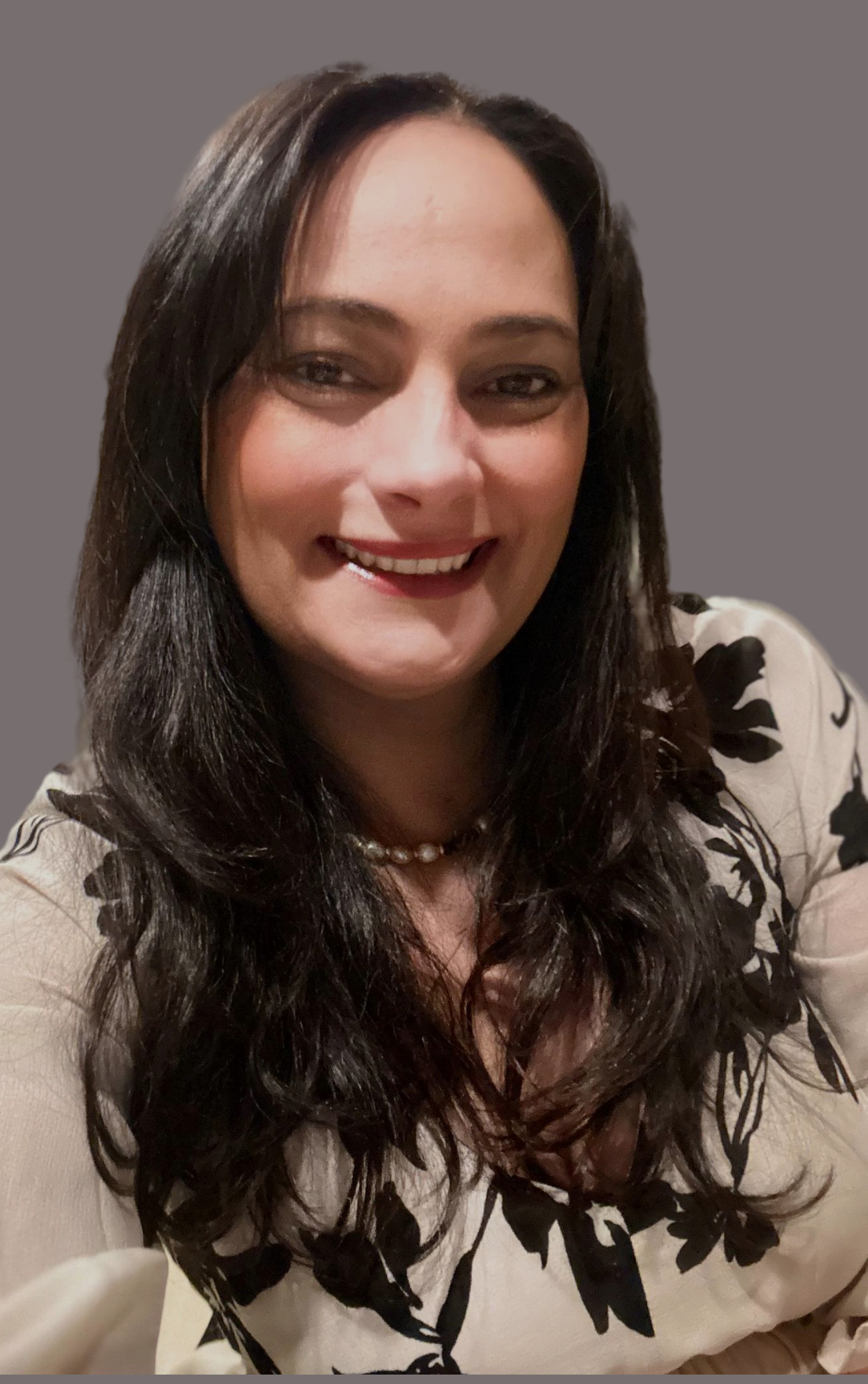 A woman with long dark hair smiling and wearing a white top with black floral patterns, a pearl necklace, and makeup, against a plain gray background.