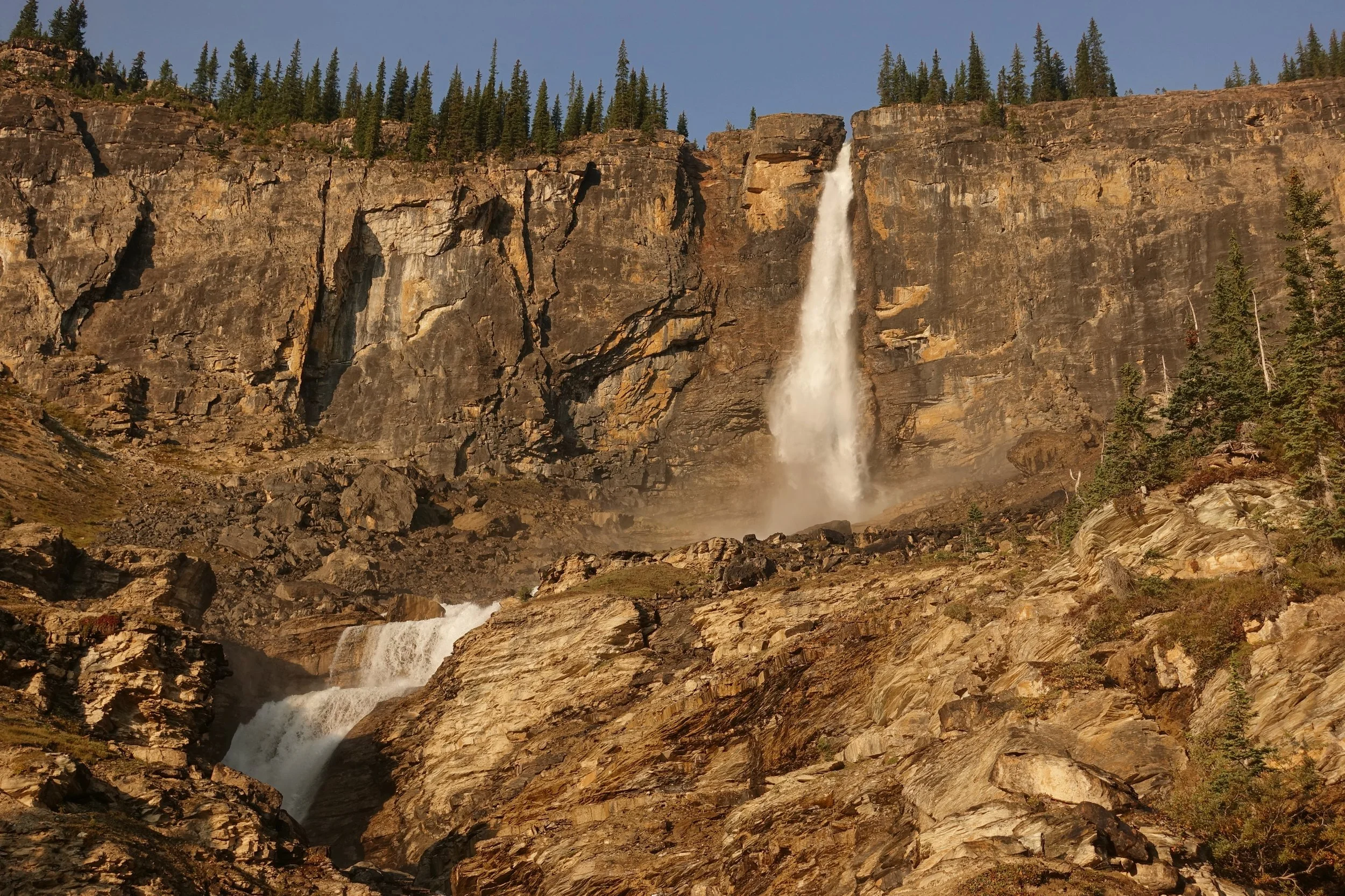 A tall waterfall flowing down a rocky cliff in a forested mountain landscape.
