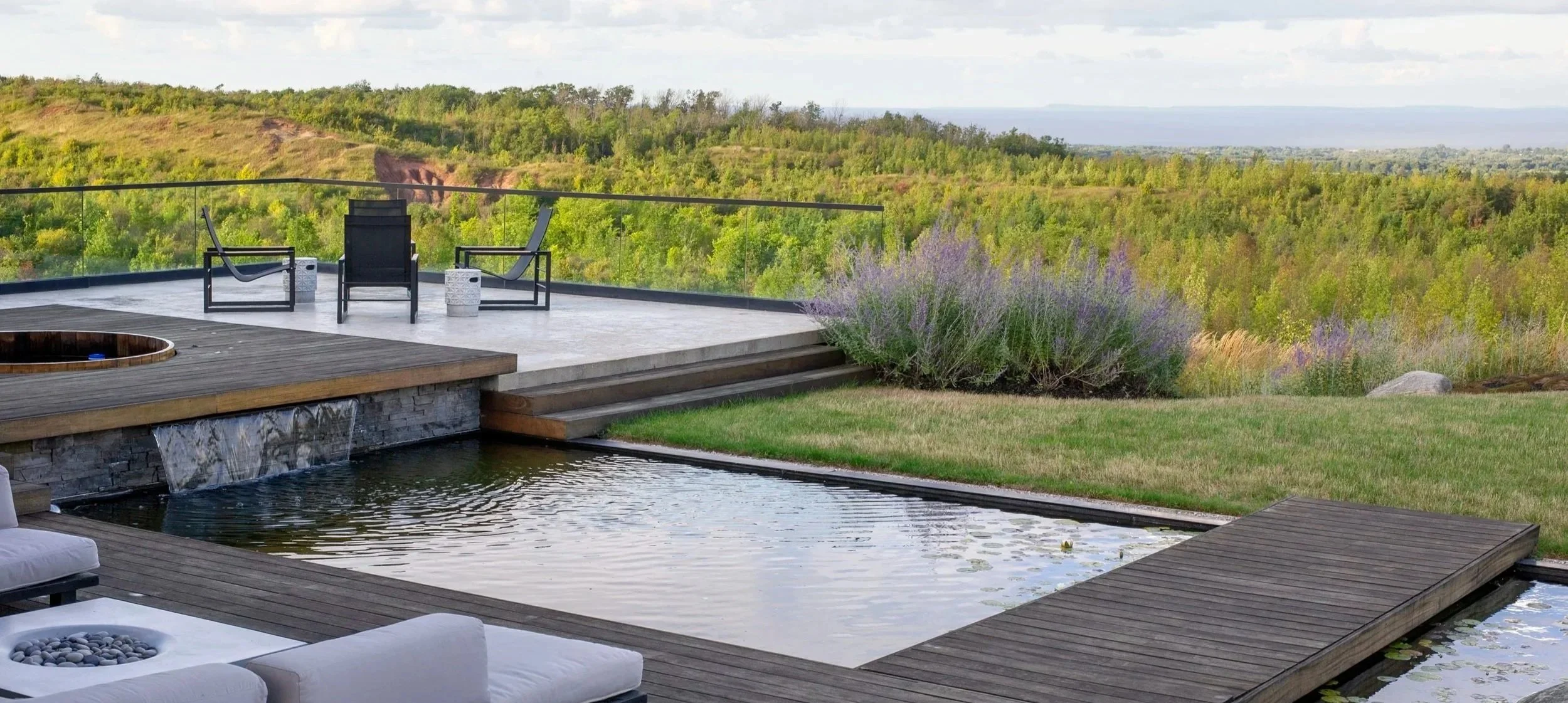 An outdoor patio features a pond with a waterfall, surrounded by wooden decking and seating, overlooking a lush green landscape with trees and hills.