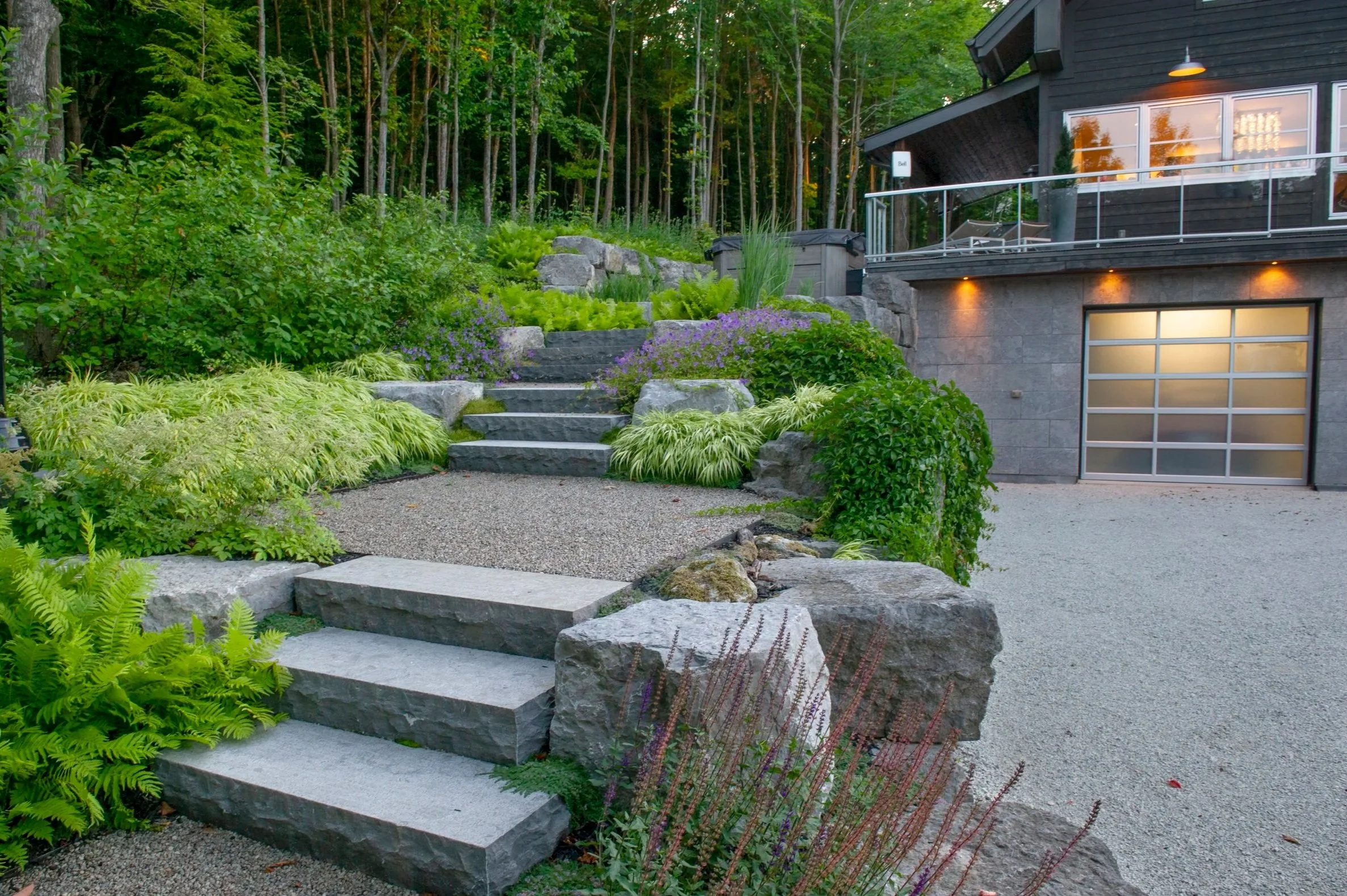 Landscaped backyard with stone steps, lush green plants, and trees, adjacent to a modern house with a garage and a balcony.