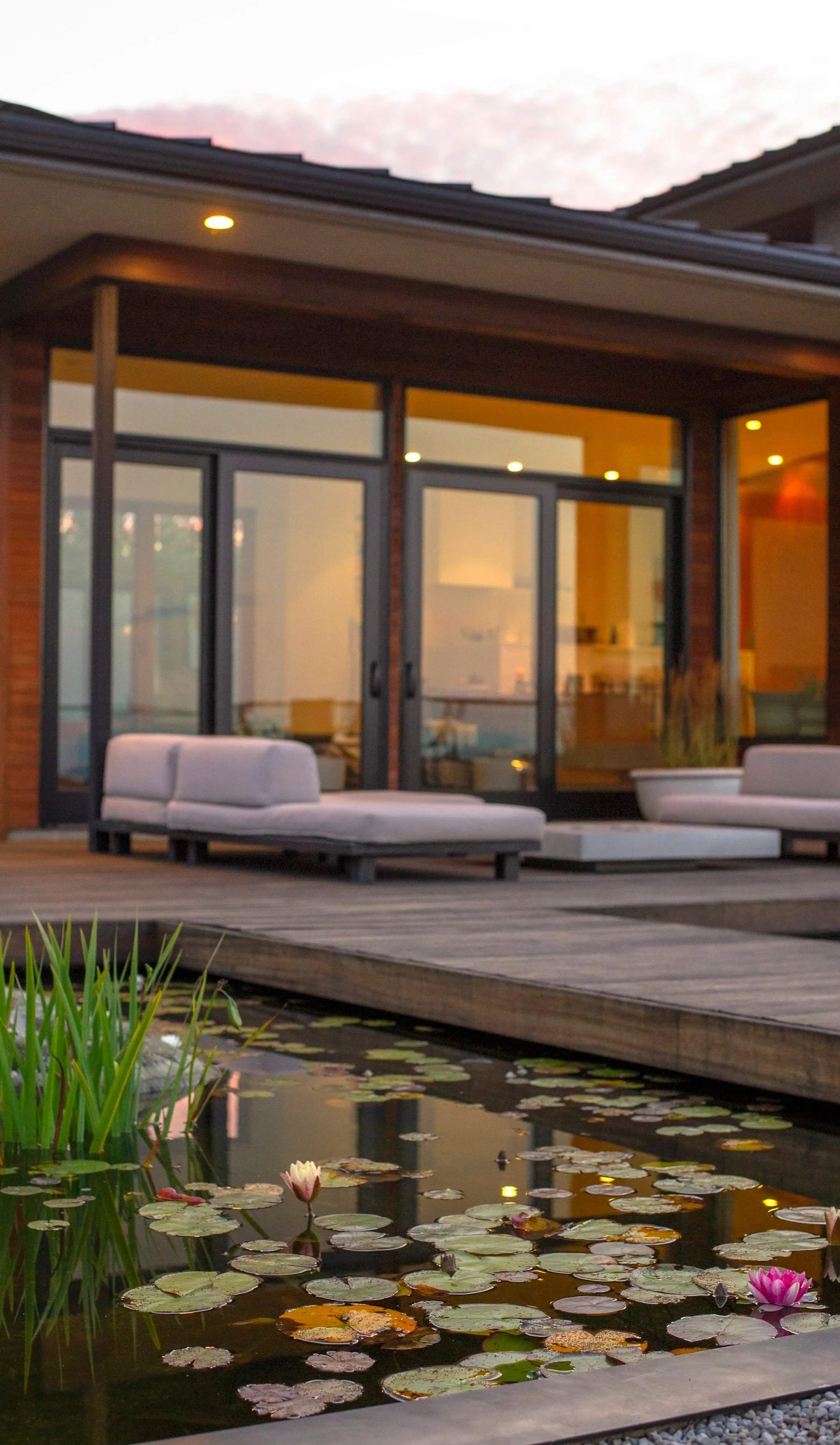 A modern house with large glass sliding doors, a wooden deck with outdoor lounge chairs, and a pond with water lilies in the foreground.