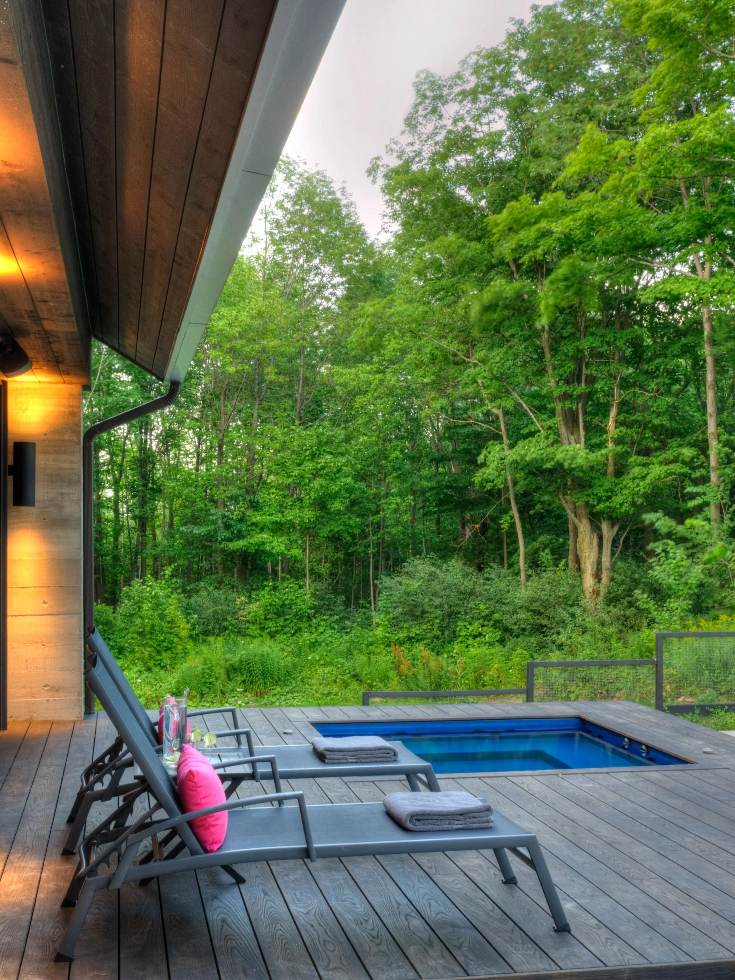 An outdoor wooden deck with two lounge chairs, towels, and a small pool, surrounded by lush green trees.