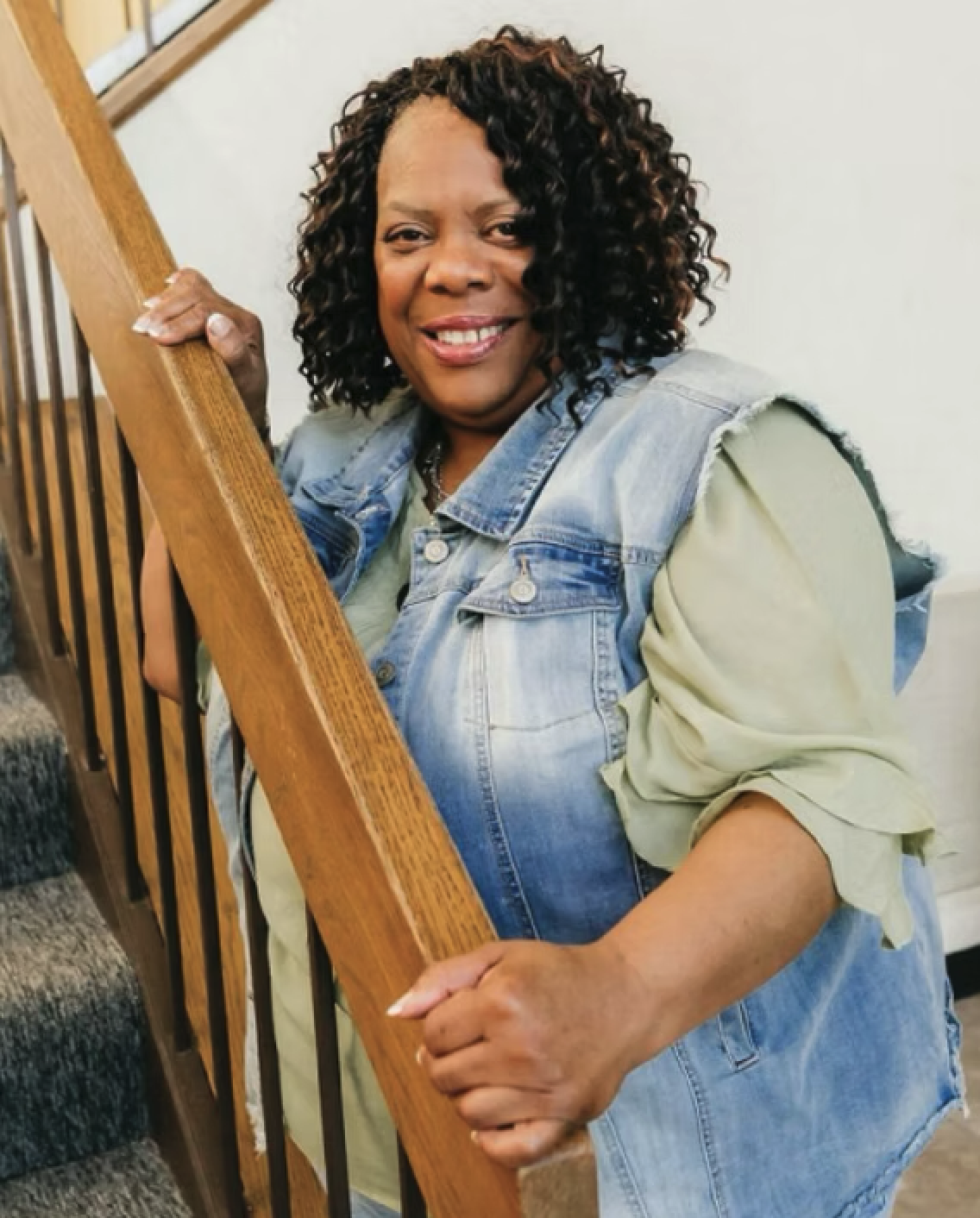 A woman with curly hair smiling and holding a wooden staircase railing.