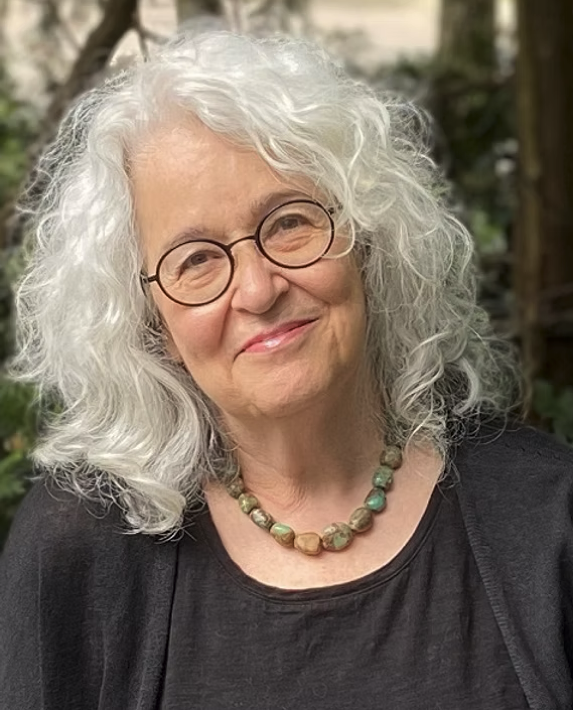 Portrait of an elderly woman with curly white hair and round glasses, smiling, wearing a black top and a beaded necklace, outdoors with greenery in the background.