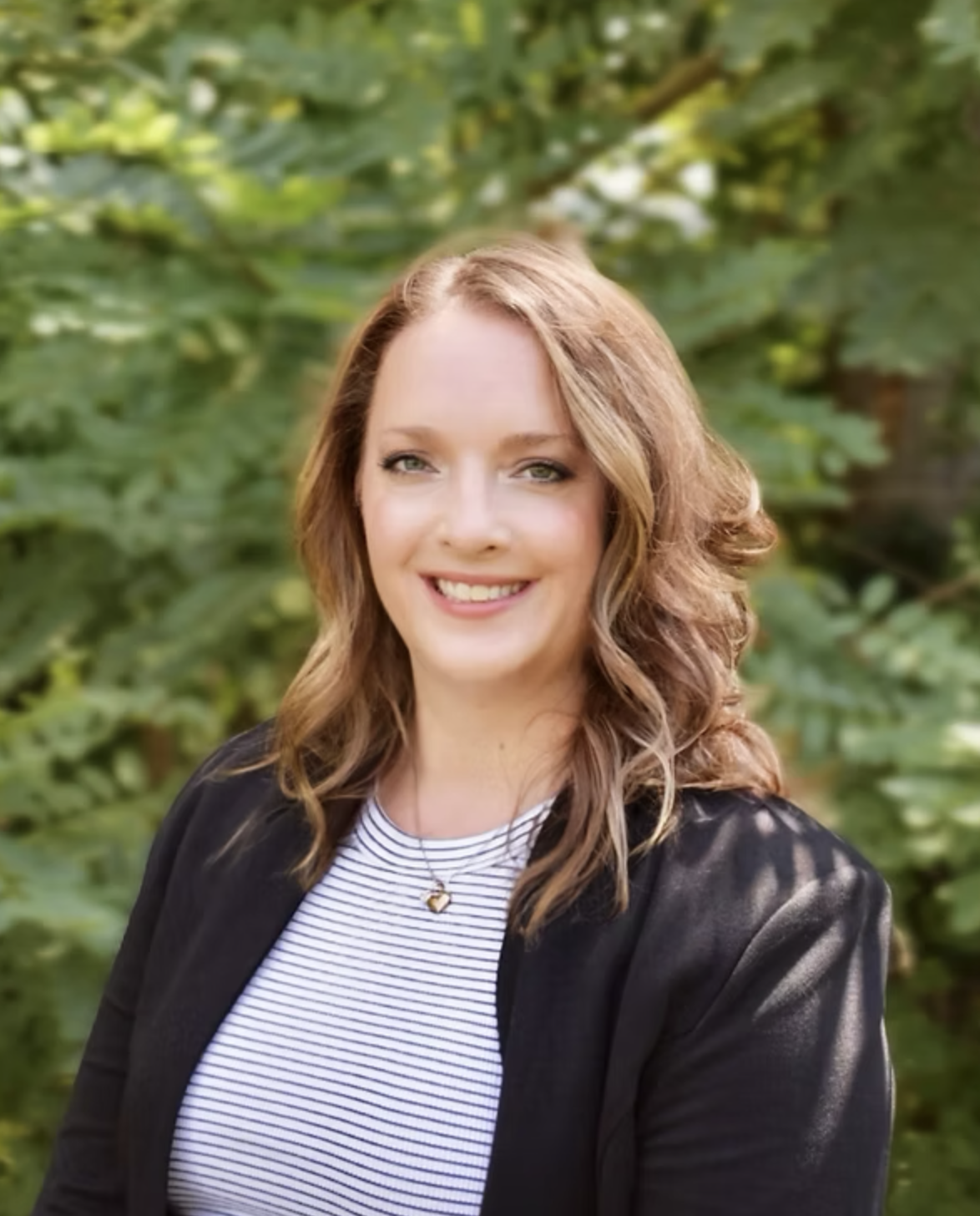 A woman with wavy, shoulder-length light brown hair, smiling, in front of green leafy trees, wearing a black blazer over a striped top and a small pendant necklace.