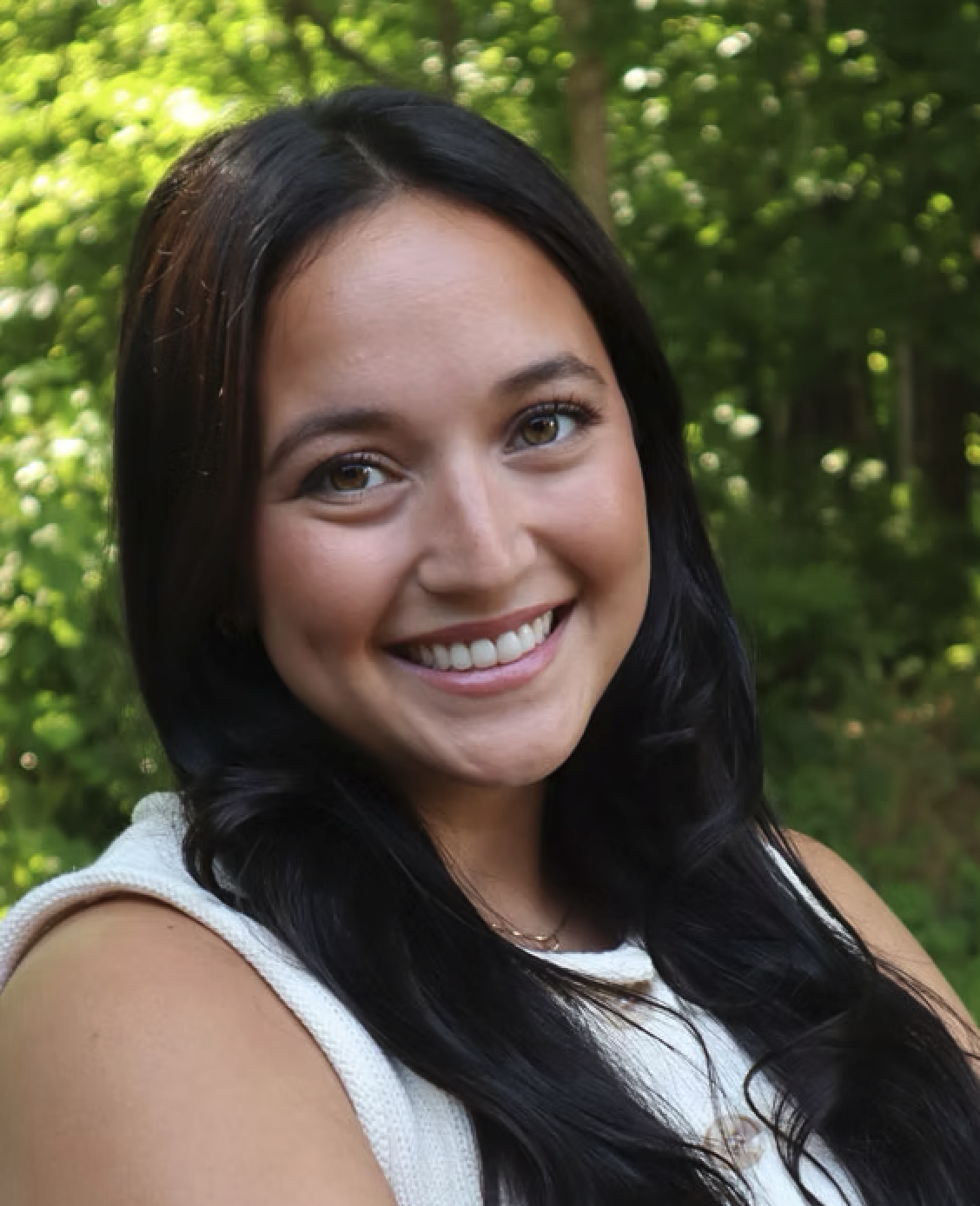 Smiling woman with black hair and light brown eyes, outdoors with green trees in the background.