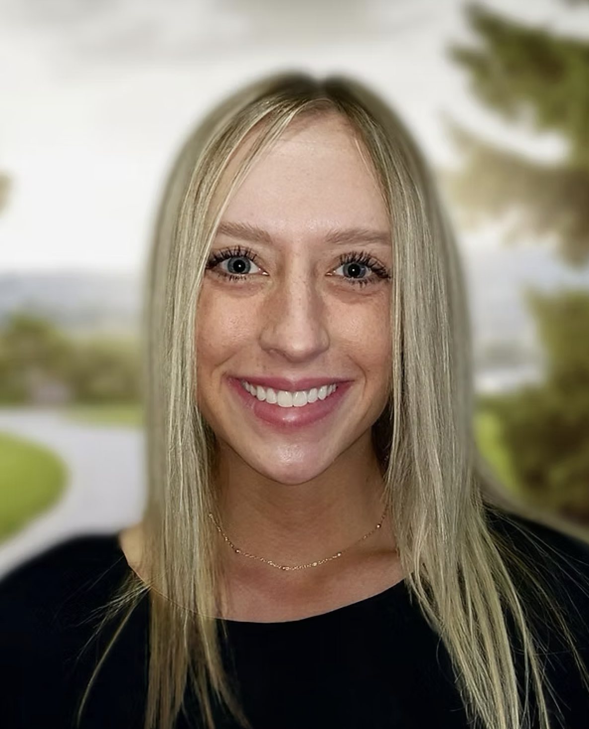 A woman with long blonde hair and blue eyes smiling outdoors with a blurred green landscape background.