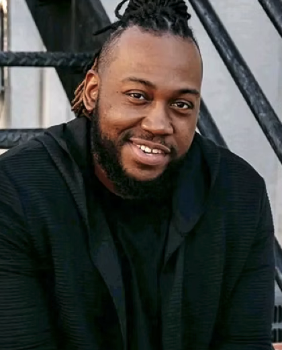 A smiling man with a beard and dreadlocks in a bun, wearing a black shirt, sitting beside metal stairs.