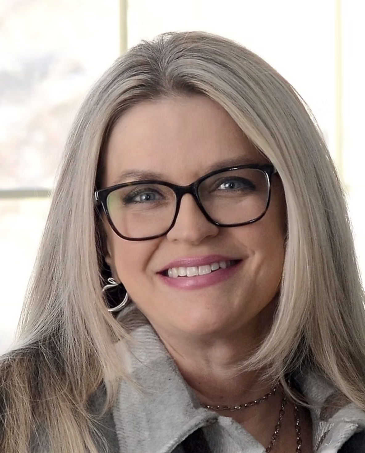 Close-up of a woman with long blonde hair, wearing glasses, smiling, and wearing earrings and a necklace.
