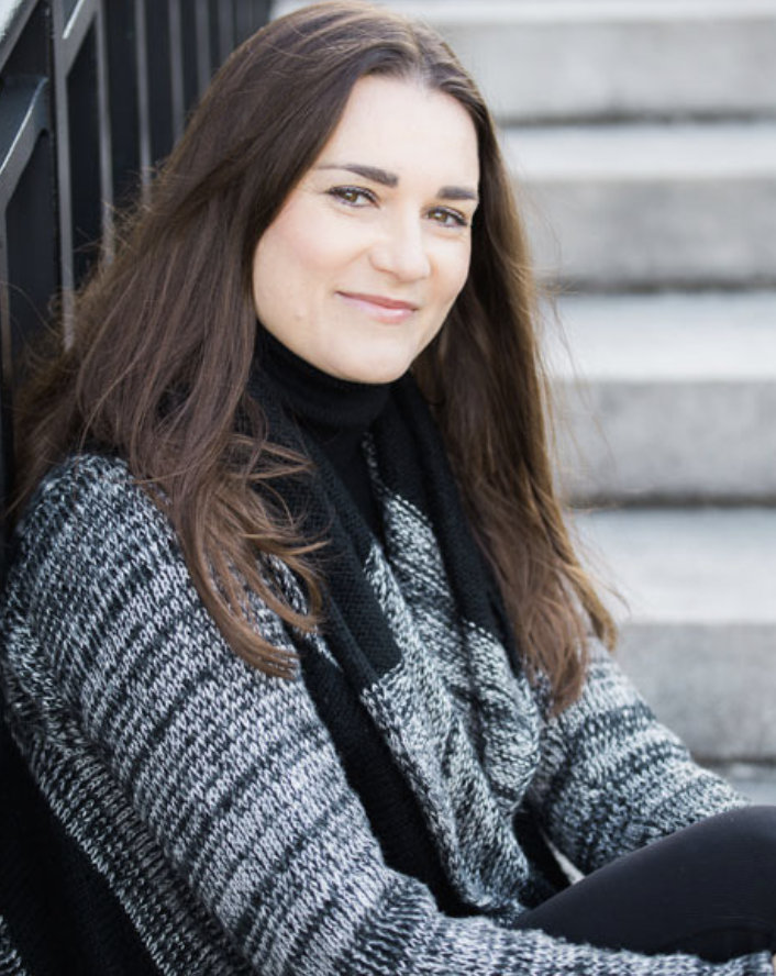 A woman with long brown hair sitting outdoors on stairs near a black metal railing, wearing a black and gray patterned sweater and a black scarf.