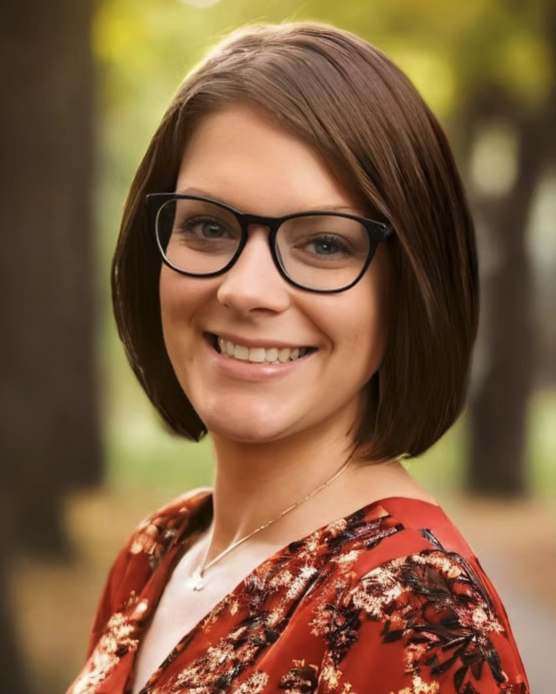 A woman with short brown hair wearing glasses, a red floral dress, and a necklace, smiling outdoors with blurred trees in the background.