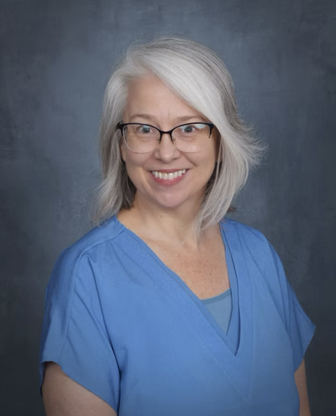 A middle-aged woman with gray hair, glasses, and a bright smile, wearing blue medical scrubs, against a dark gray background.