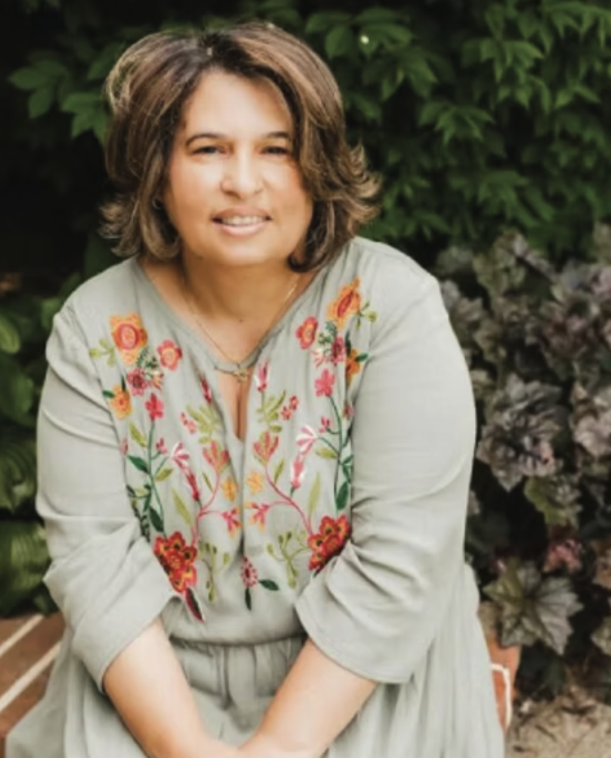 A woman with shoulder-length brown hair, wearing a light-colored dress with colorful floral embroidery, sitting outdoors in front of green foliage and purple plants.