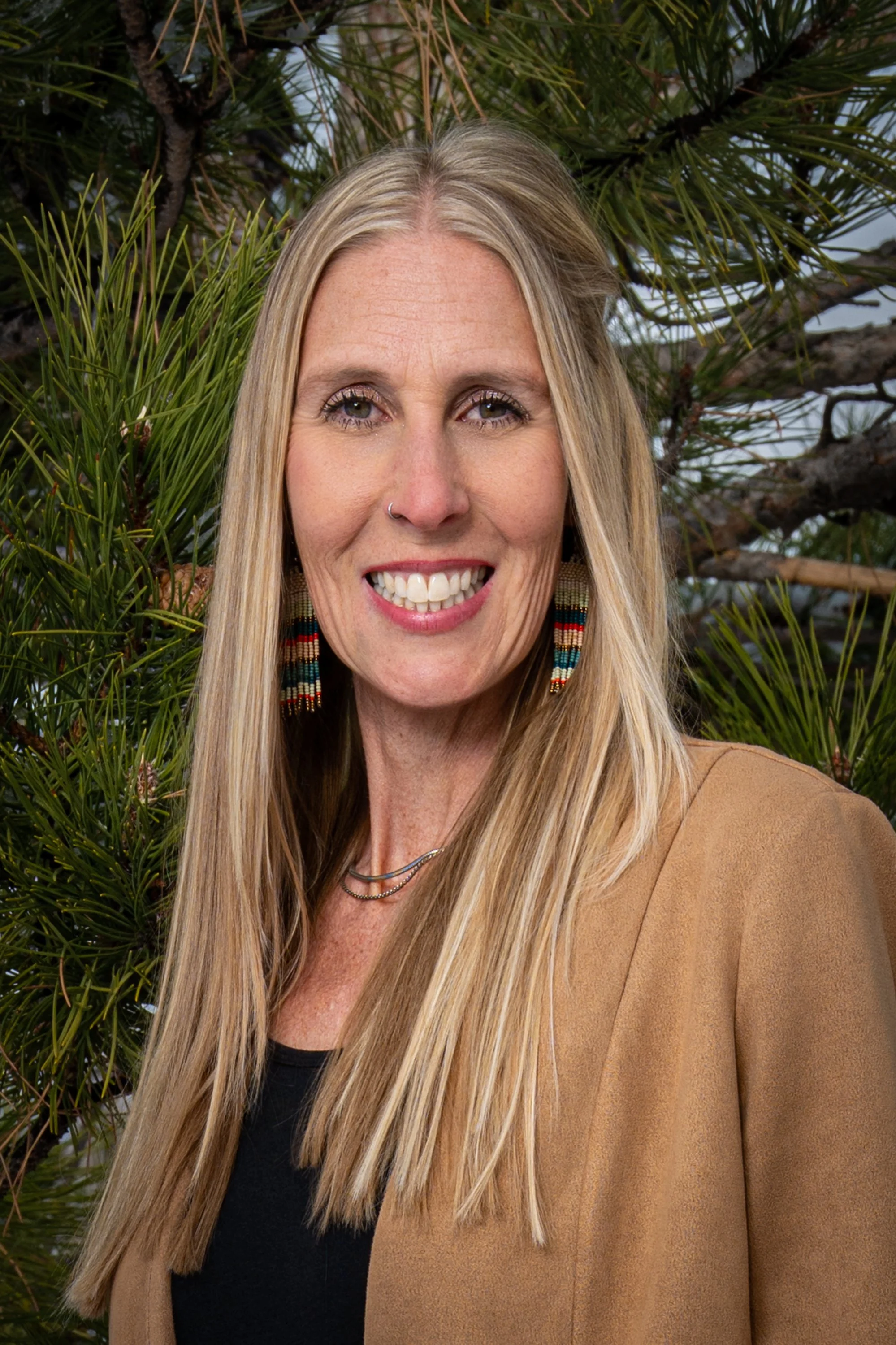 Julie Smith, a woman with long blonde hair, wearing beaded earrings, a necklace, a black top, and a tan blazer, standing in front of green pine tree branches outdoors.