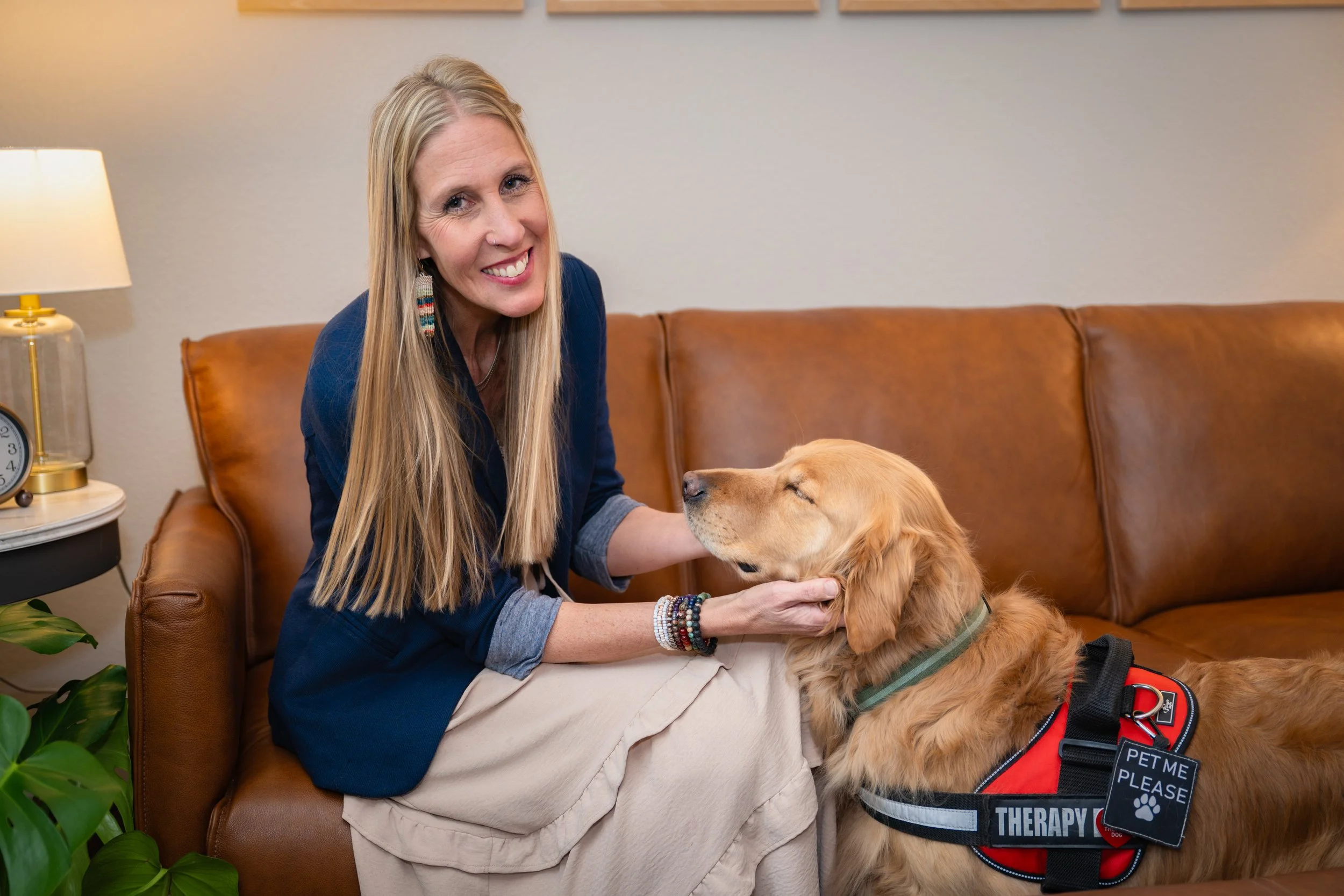 Julie Smith smiling as she pets a golden retriever therapy dog (River) on a tan leather couch in her cozy office.