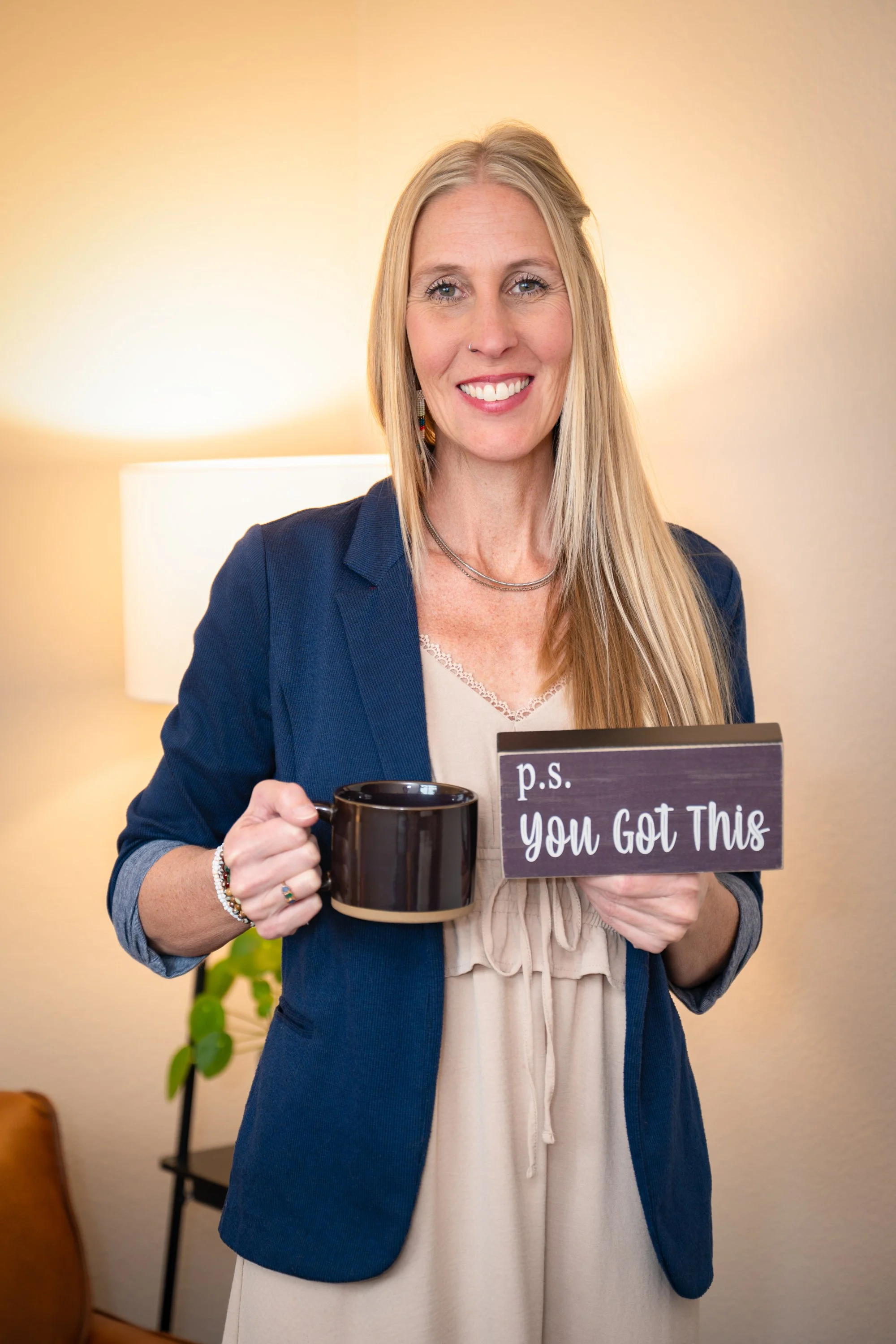 Julie Smith smiling holding a black mug and a sign that reads 'p.s. You Got This' in her cozy office with a lamp and a plant in the background.