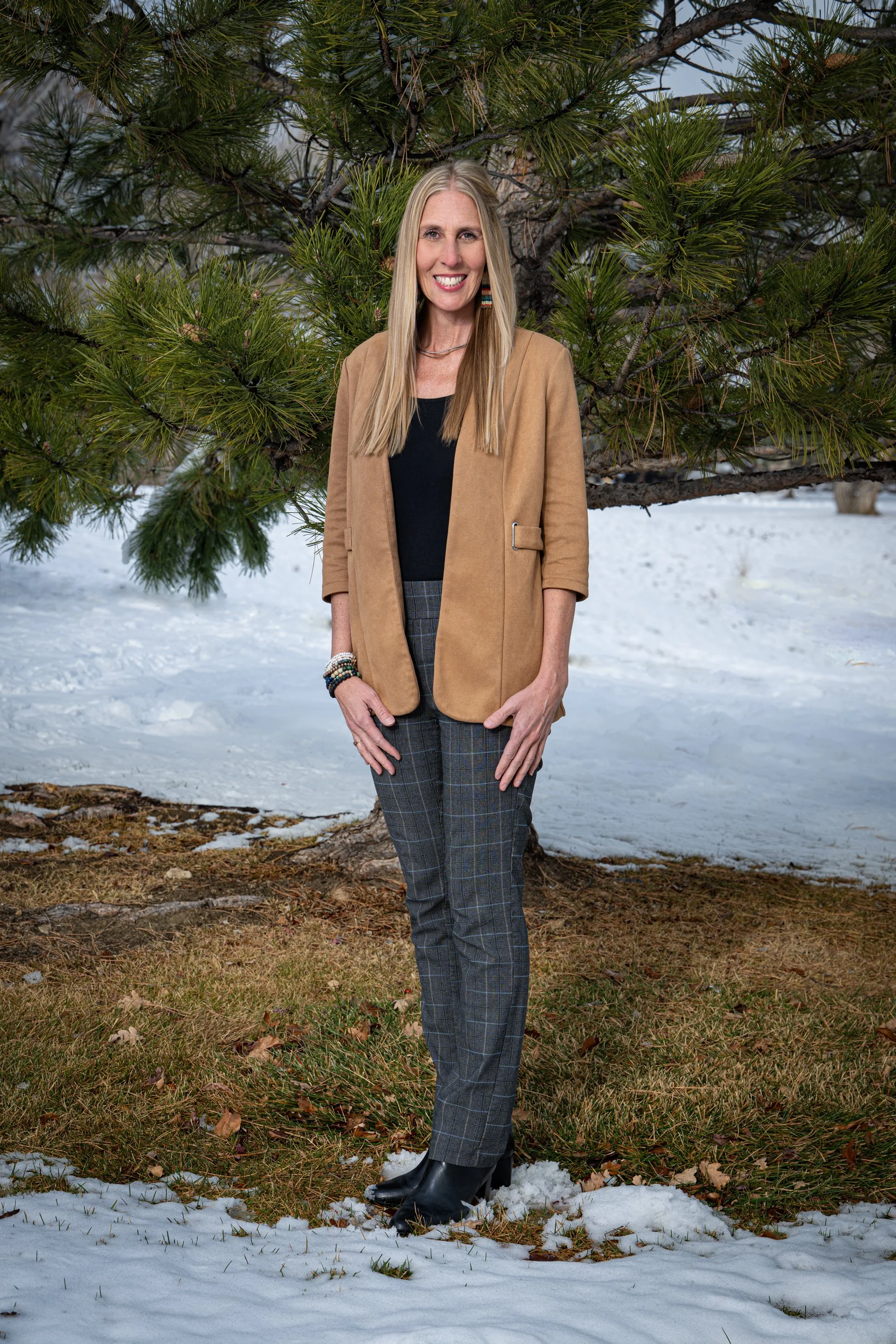 Julie Smith standing outdoors on snow, smiling, with a pine tree behind her, wearing a tan blazer, black top, plaid pants, and black boots.