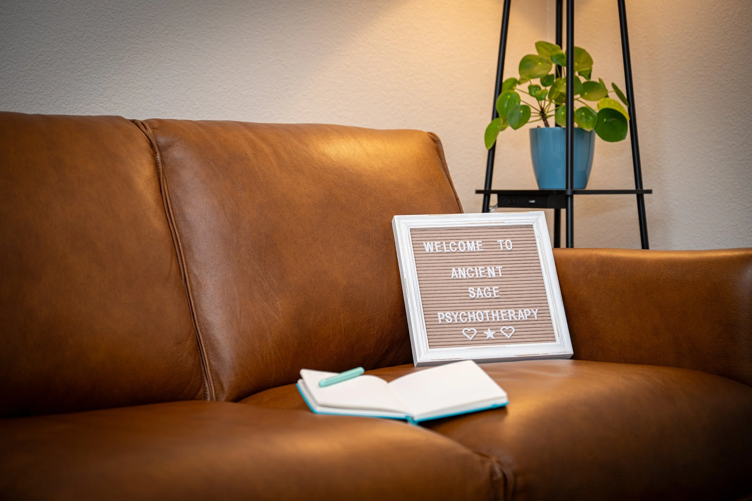Julie Smith's brown leather office couch with an open notebook and a pen on it, a sign that says "Welcome to Ancient Sage Psychotherapy," and a potted plant on a black metal stand in the background.