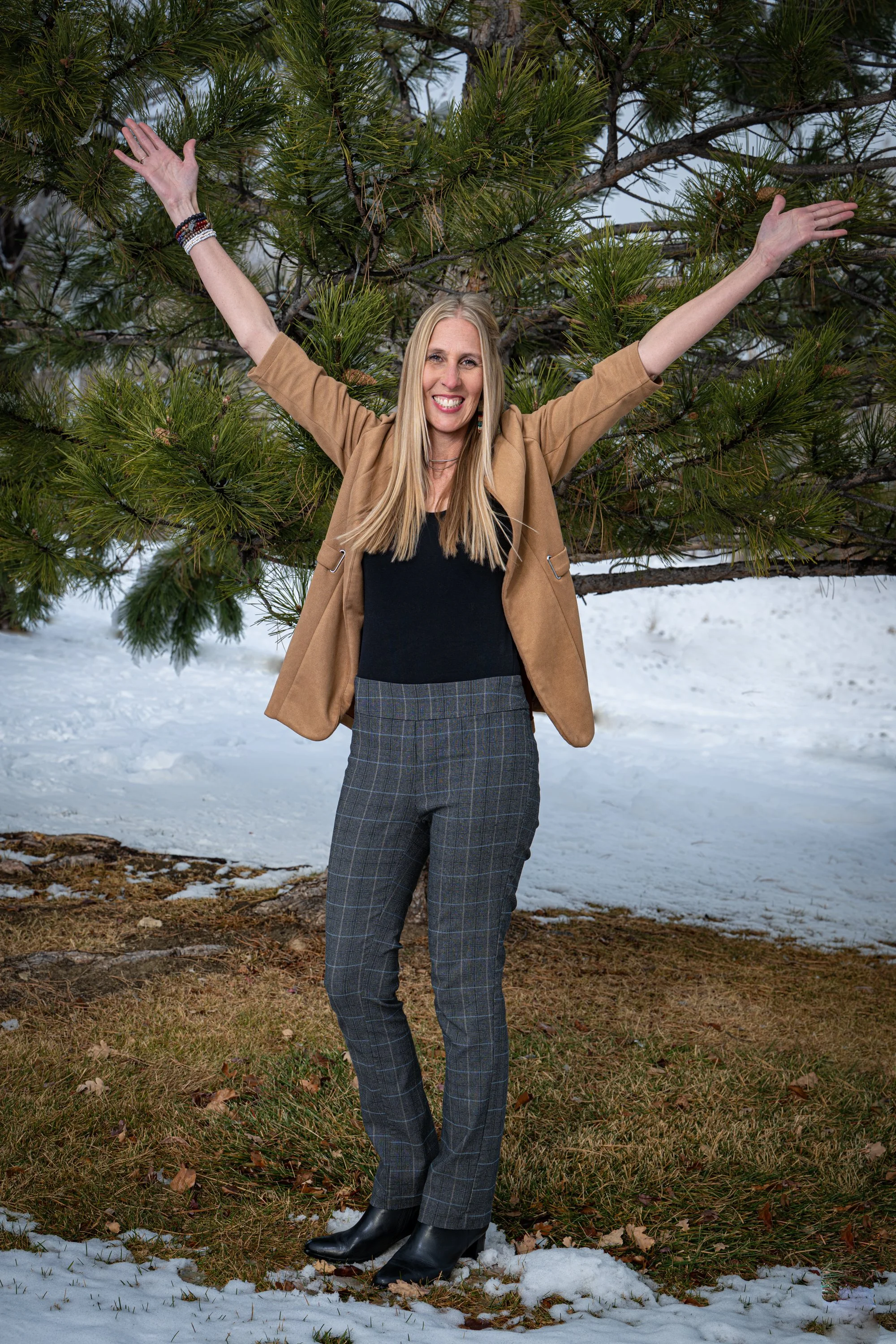 Julie Smith standing outdoors on a patch of grass and snow, smiling with arms raised, in front of a pine tree.