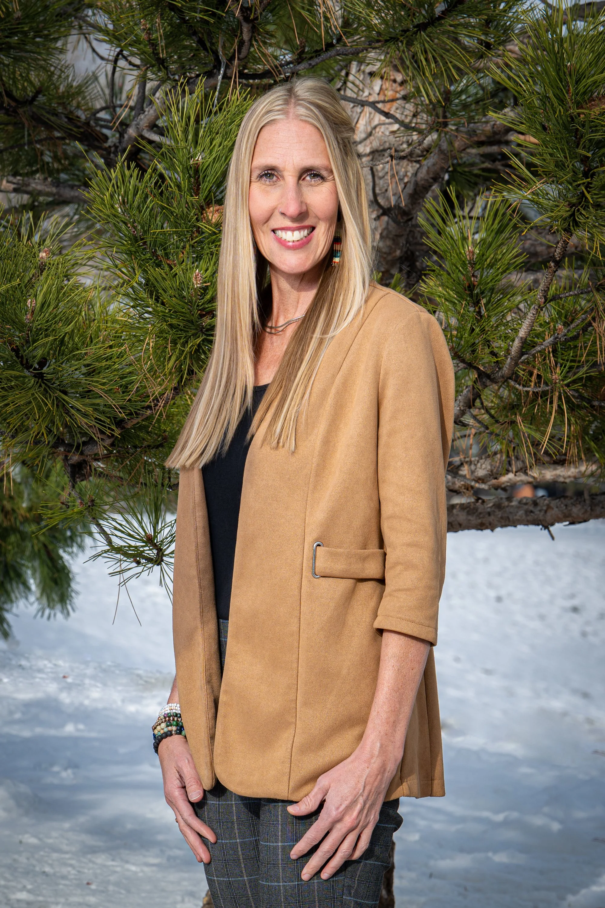 Julie Smith smiling, standing outdoors near a pine tree with snow on the ground, wearing a tan jacket, black top, and plaid pants.