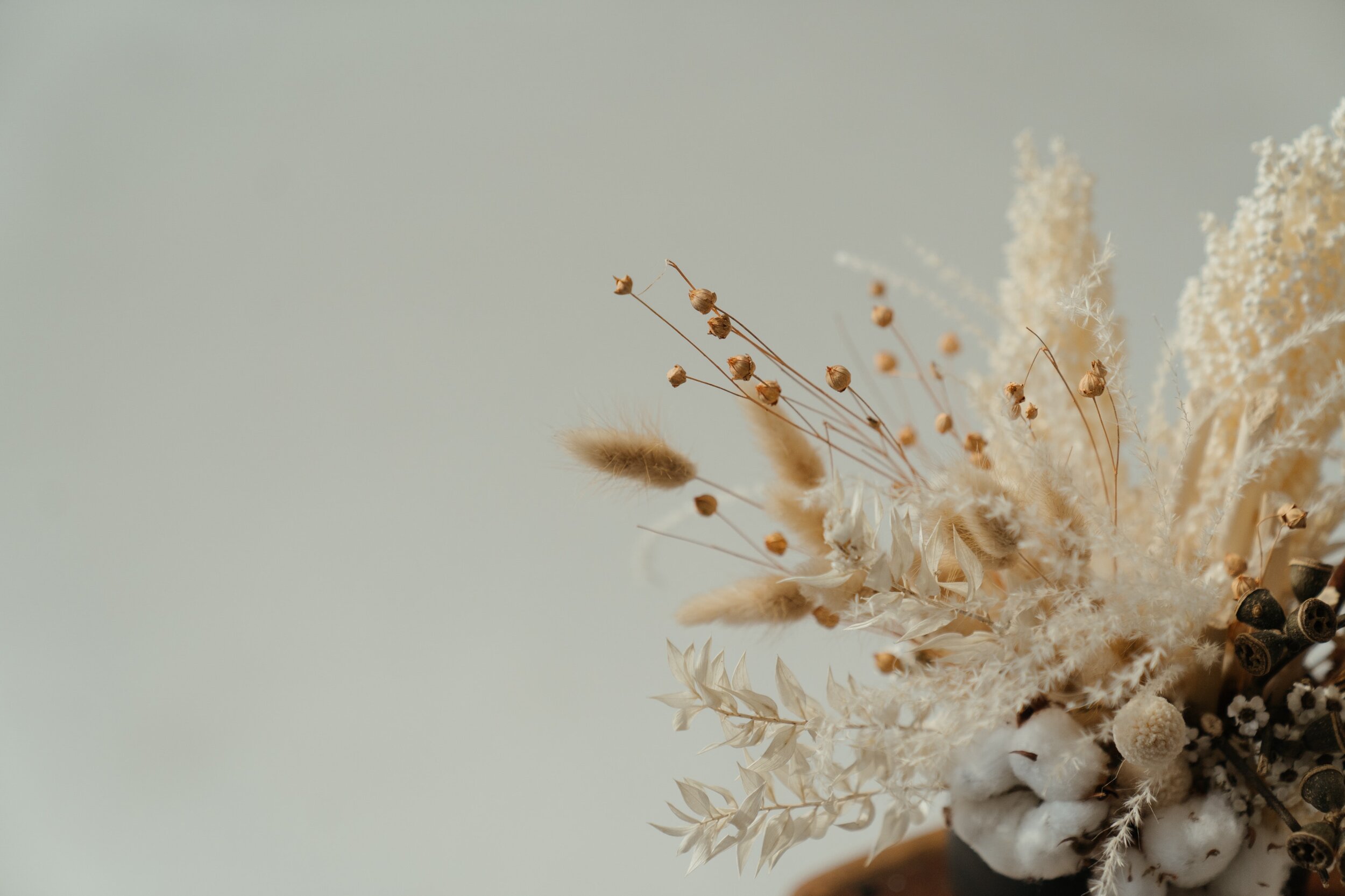 A bouquet of dried flowers and grasses in neutral tones, including fluffy white cotton, seed pods, and various dried leaves and stems, arranged in a brown vase.