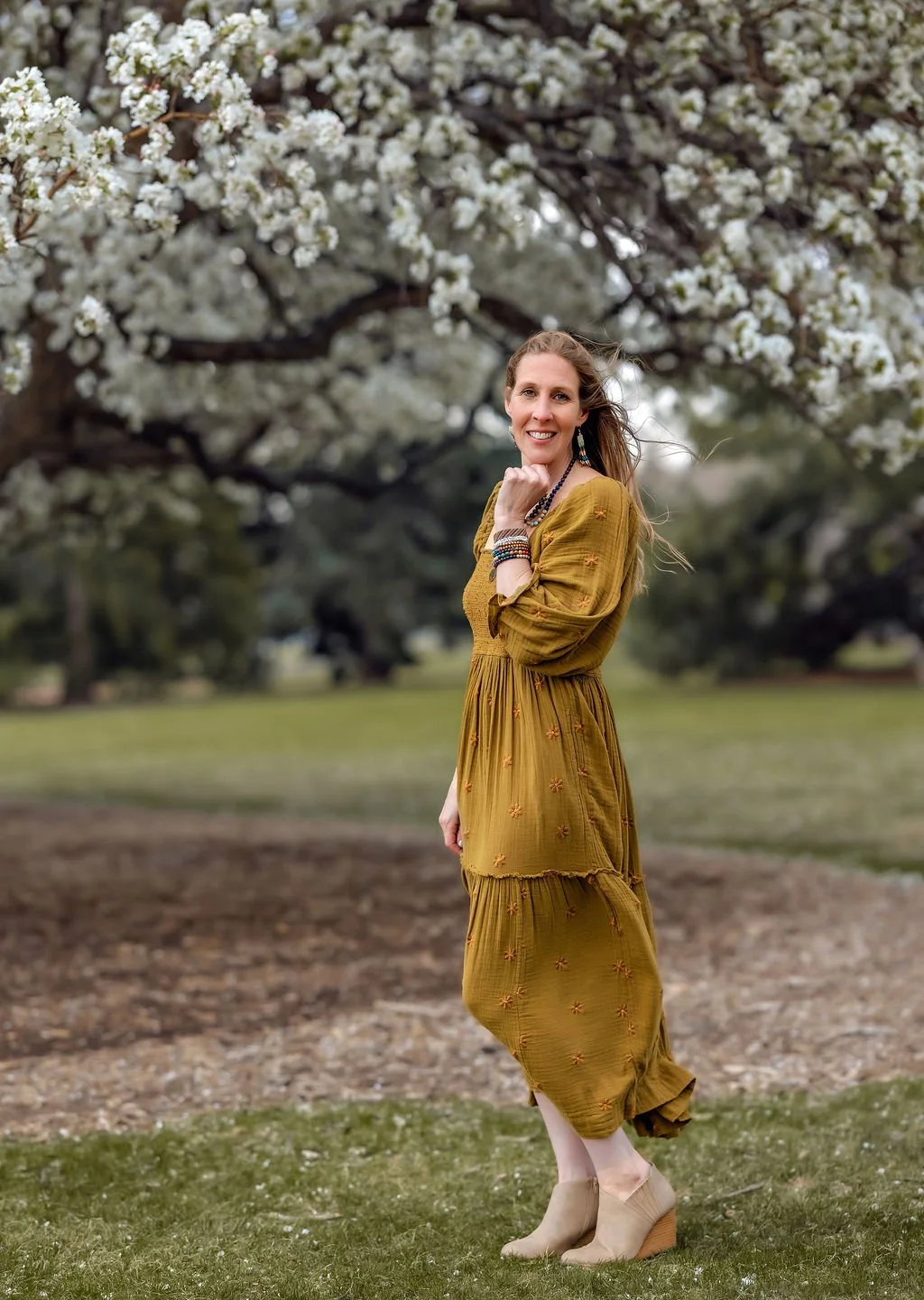 A woman in a mustard yellow dress with embroidered flowers standing outdoors near a blooming tree with white flowers, on a grassy and dirt path, smiling at the camera.