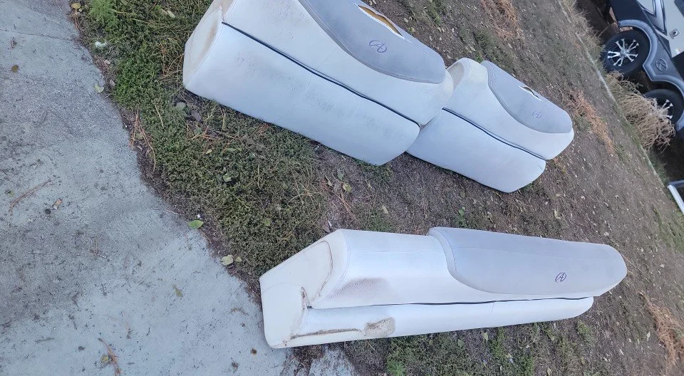 White automotive seats placed on the ground outdoors near a dirt and grass area next to a sidewalk.