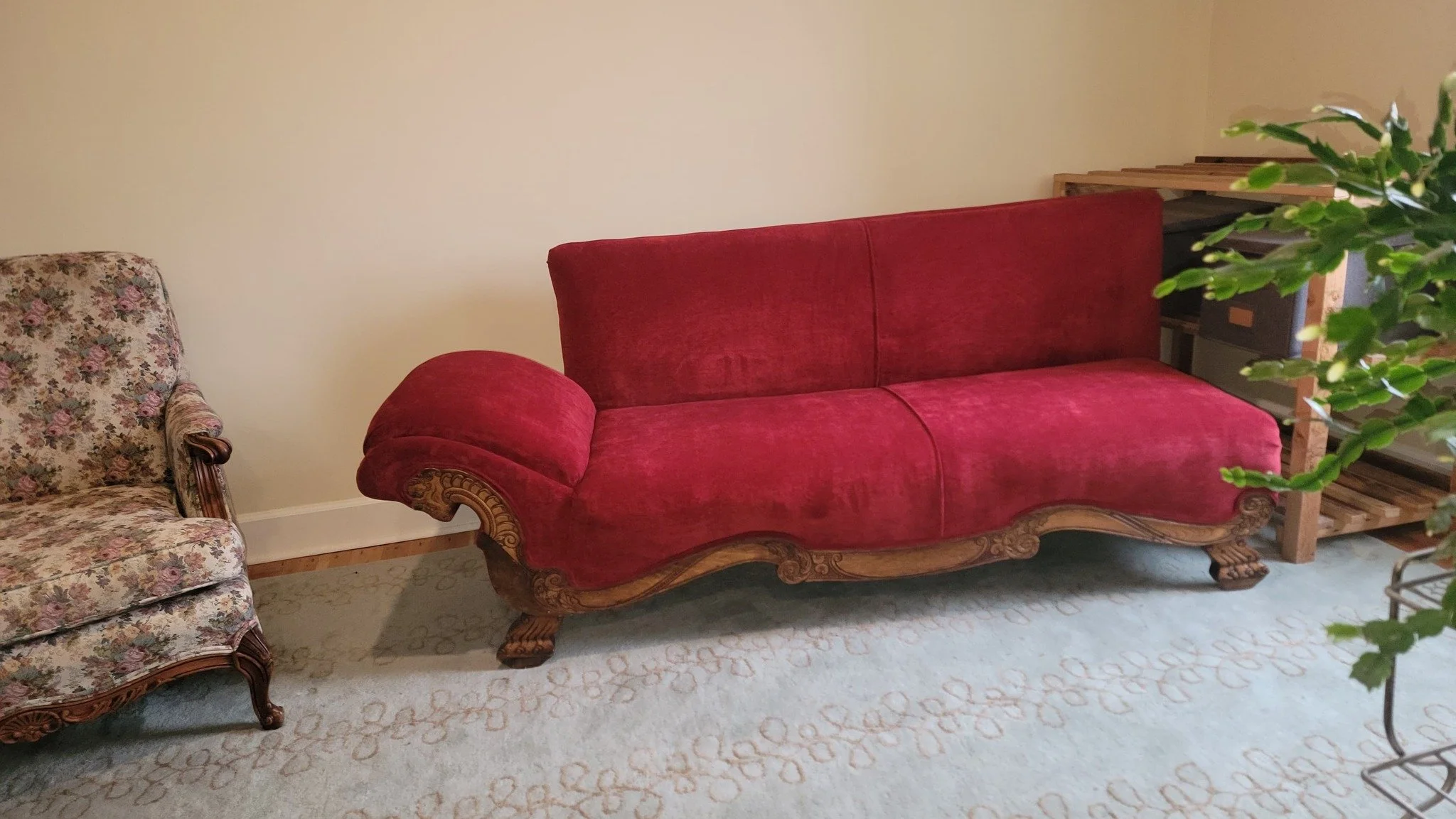 A vintage red velvet sofa with carved wooden legs and trim, positioned against a cream-colored wall, next to a floral upholstered armchair and a wooden shelving unit, all on a beige patterned rug.