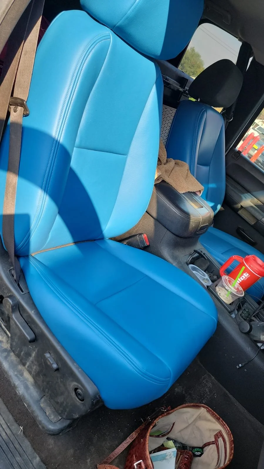 Photo of the interior of a vehicle showing the front seats with bright blue seats, a brown cloth on the center console, and various items including a red thermos, plastic cup, and a bag on the floor.