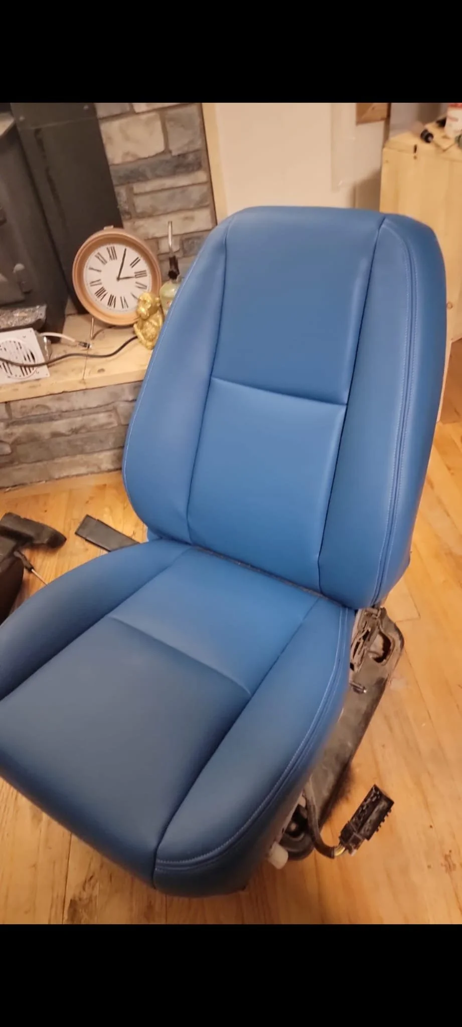 A blue car seat placed on a black platform inside a room with wooden floors, a stone fireplace, a clock, and decorative items on a stone ledge in the background.