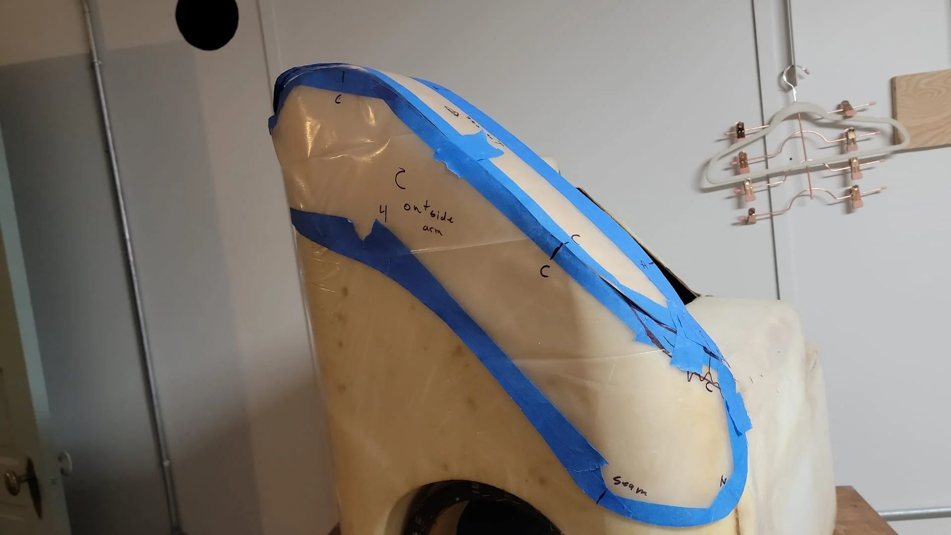 A skeleton model wrapped in blue painter's tape with handwritten notes, placed on a table in a room with a white door and a hanging clothing rack in the background.