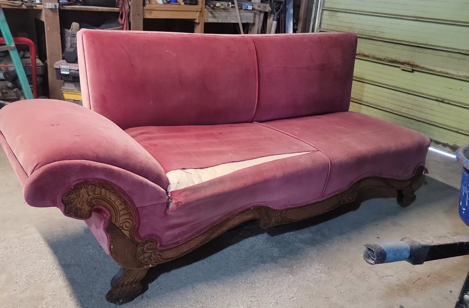 Old pink velvet sofa with carved wooden legs and trim, in a garage with cluttered shelves and tools in the background.