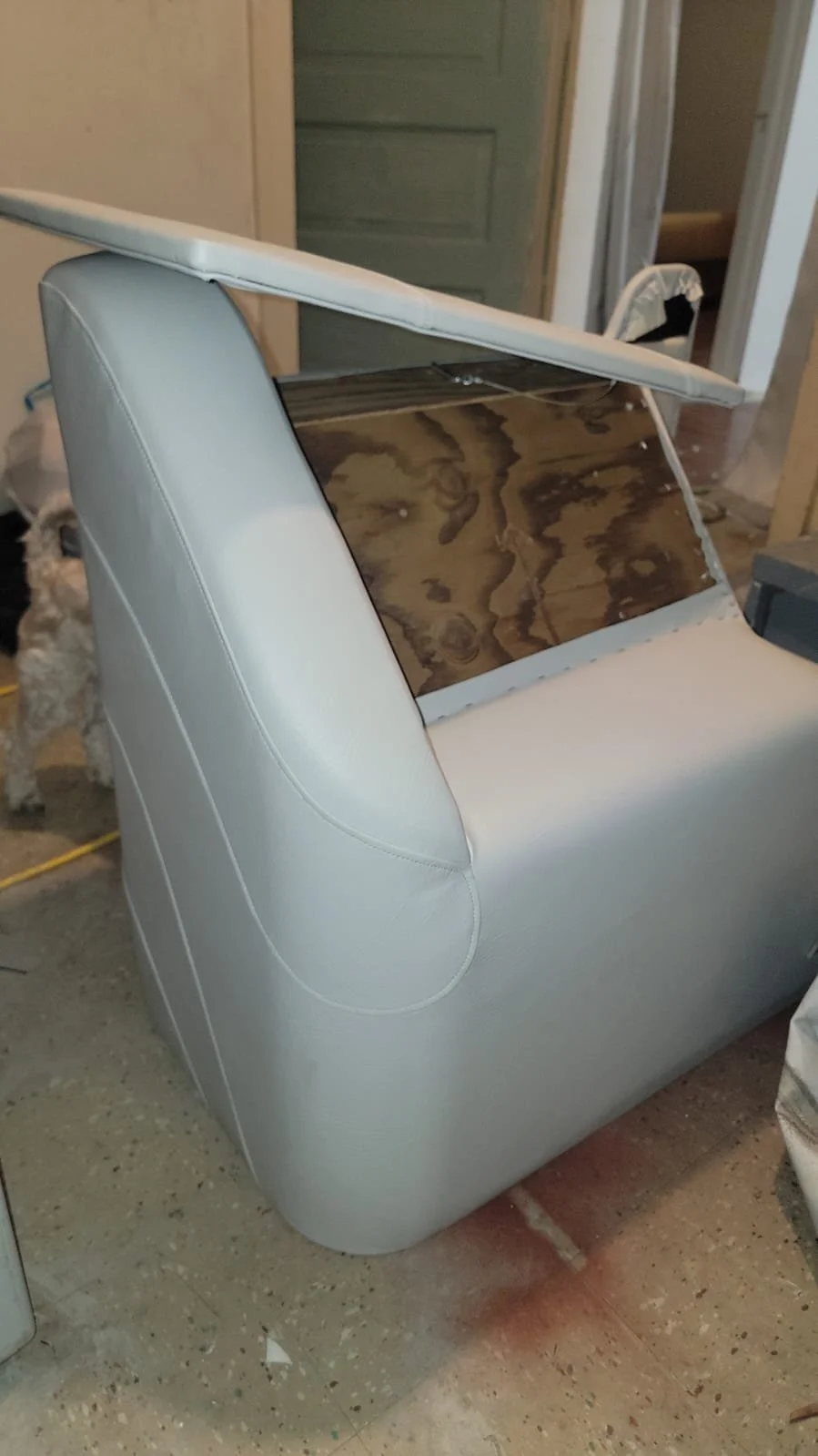 A piece of furniture with a white leather exterior, a wooden panel on top, and a cushion on the side, situated in a room with a concrete floor and a doorway in the background. A small dog is partially visible behind the furniture.