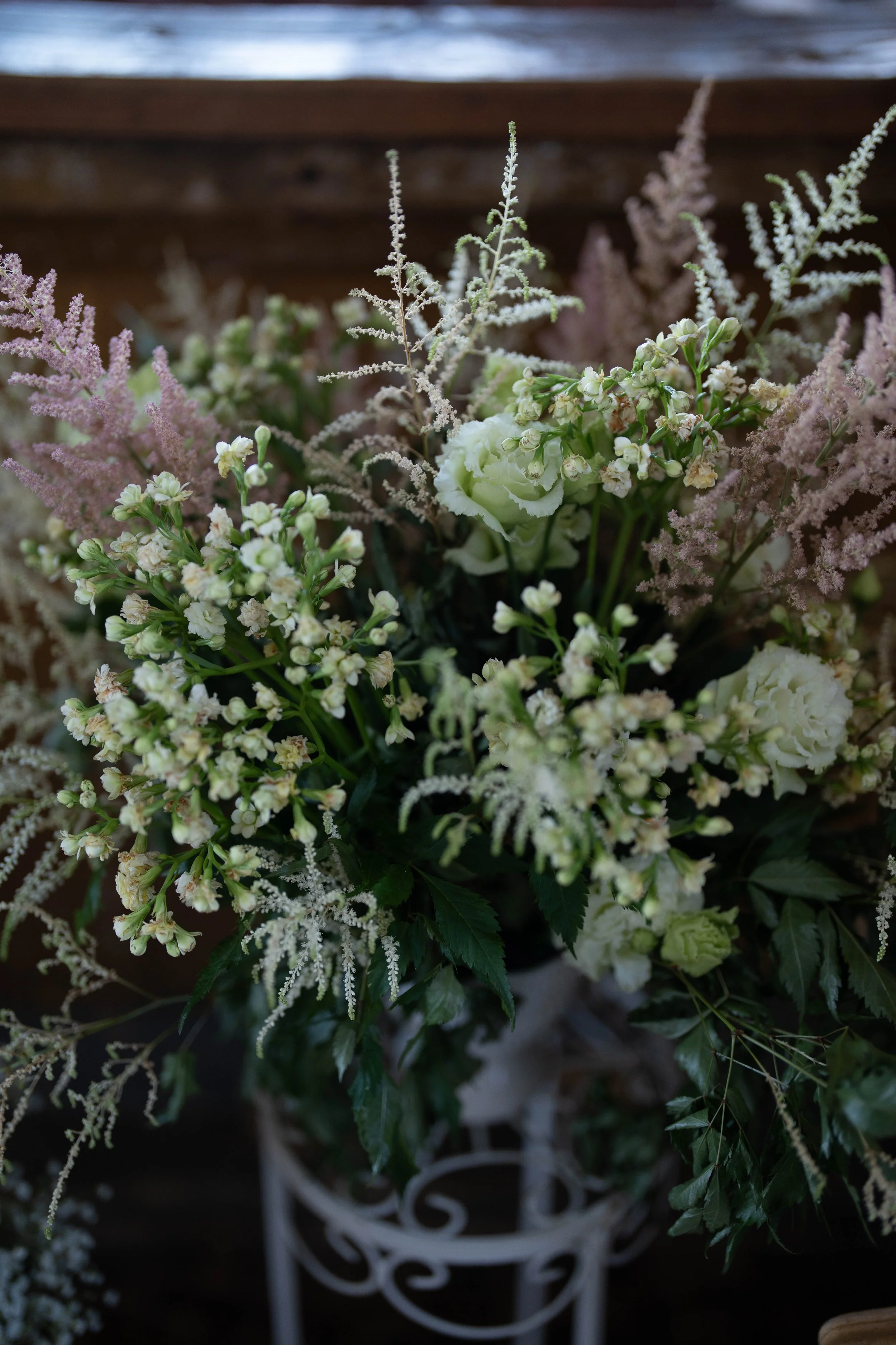 A bouquet of white and pale pink flowers with green foliage in a decorative white vase, set against a wooden background.