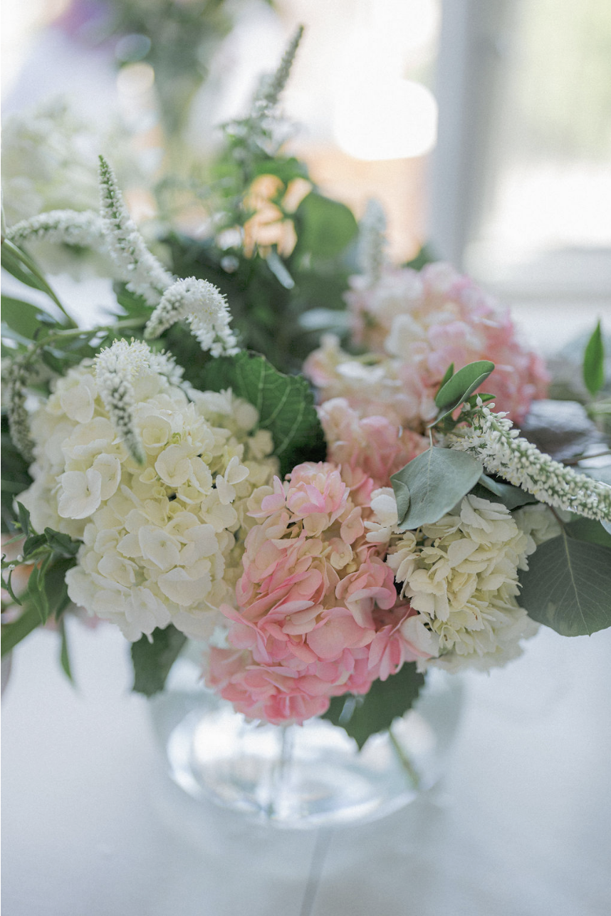 A glass vase filled with a bouquet of white, pink, and cream-colored hydrangeas and green foliage, set on a light-colored surface near a window.