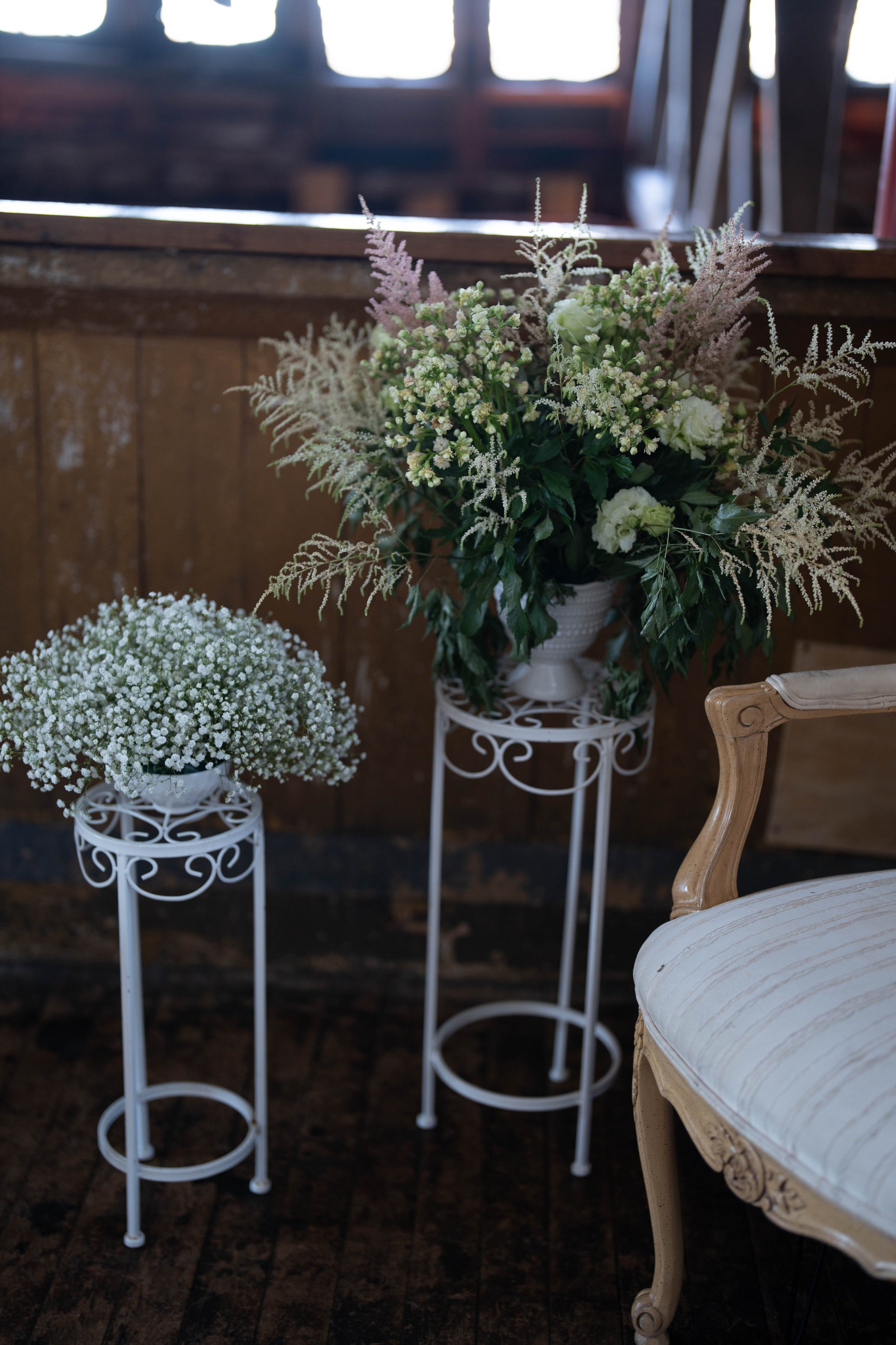 Two white metal plant stands with white flowers and lavender flowers, beside a vintage cream-colored chair with striped upholstery, set against a wooden wall, in a rustic indoor setting.