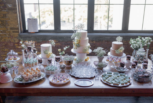 Decorative dessert table with tiered cakes, cupcakes, and sweets against a large window backdrop.