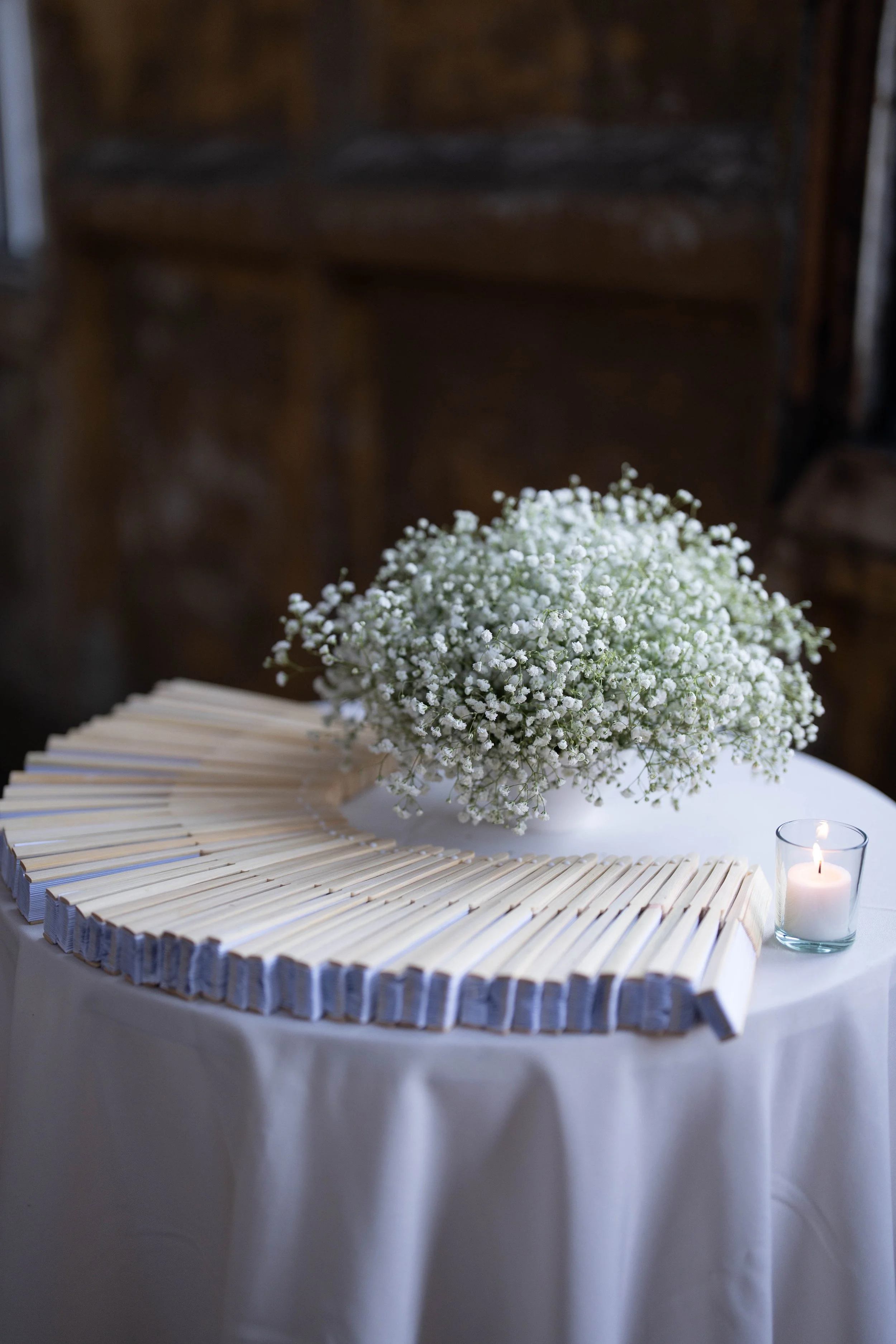 White floral arrangement of baby's breath on a white table, accompanied by a lit candle in a glass holder, with wooden vintage background.