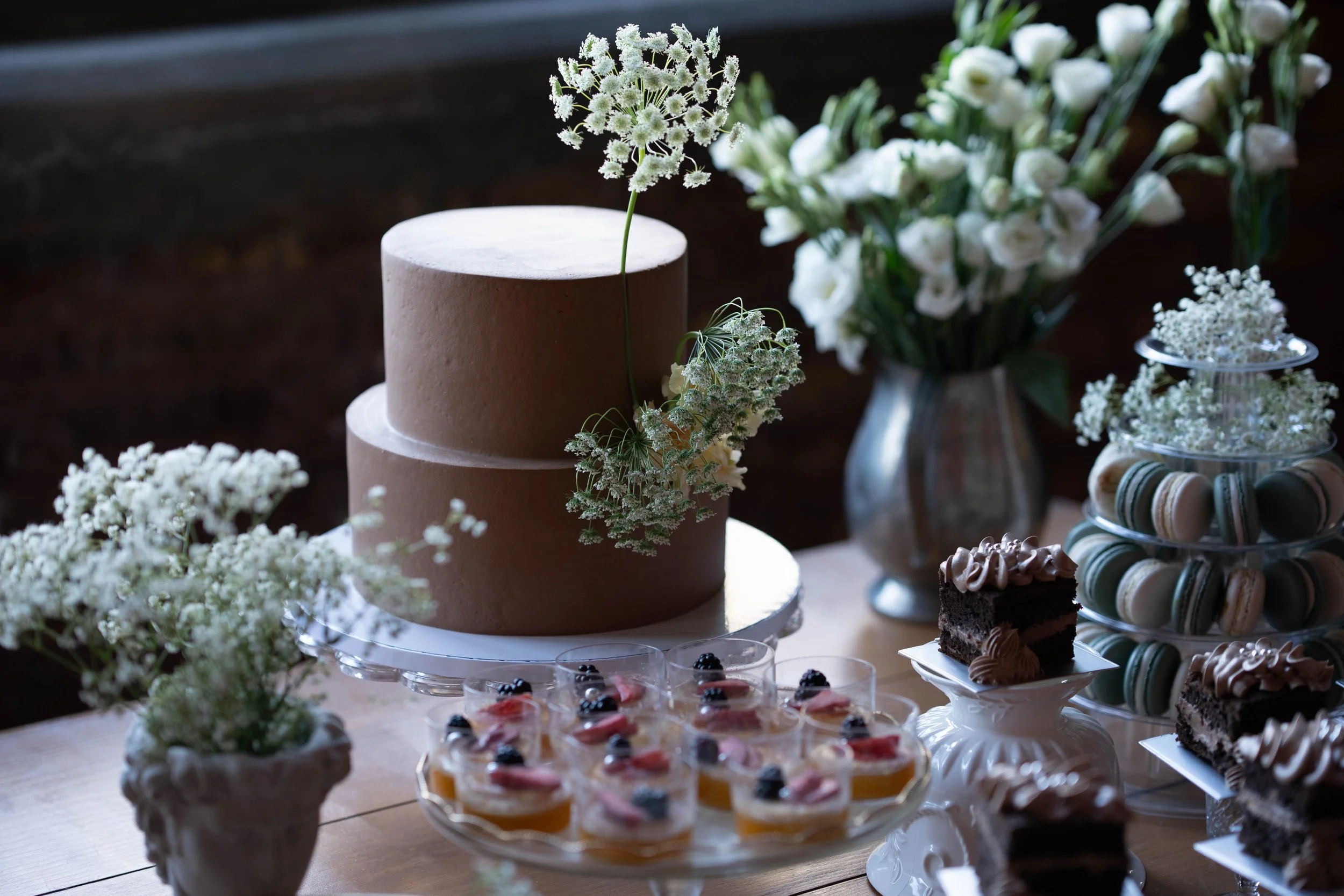 A decorated table featuring a two-tiered brown cake with white flowers, surrounded by an assortment of desserts including macarons, mini fruit tarts, and chocolate cake pieces, with white flower arrangements in vases for decoration.